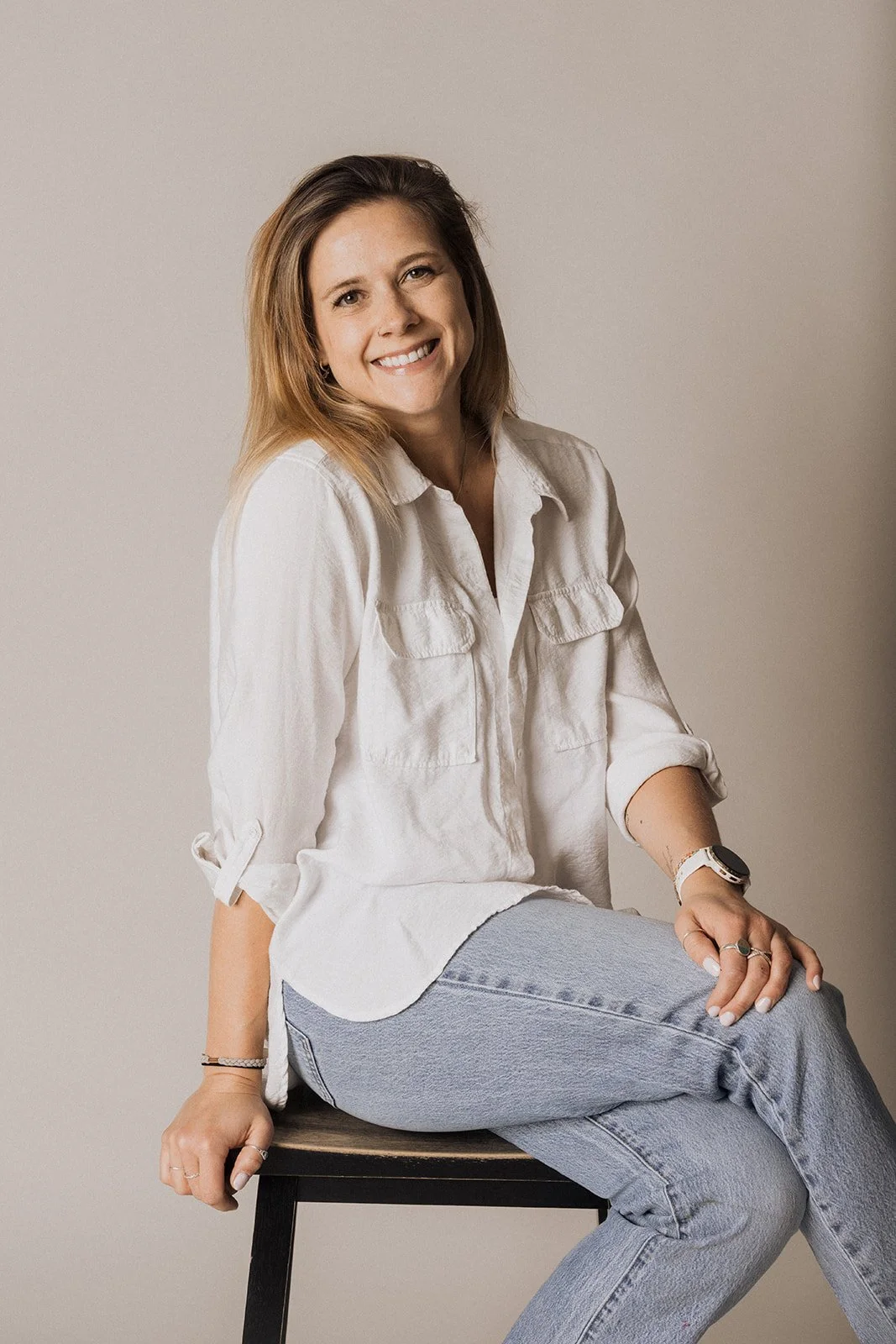 A woman with shoulder-length brown hair wearing a beige button-down shirt and light blue jeans, sitting on a black stool against a neutral background, smiling at the camera.