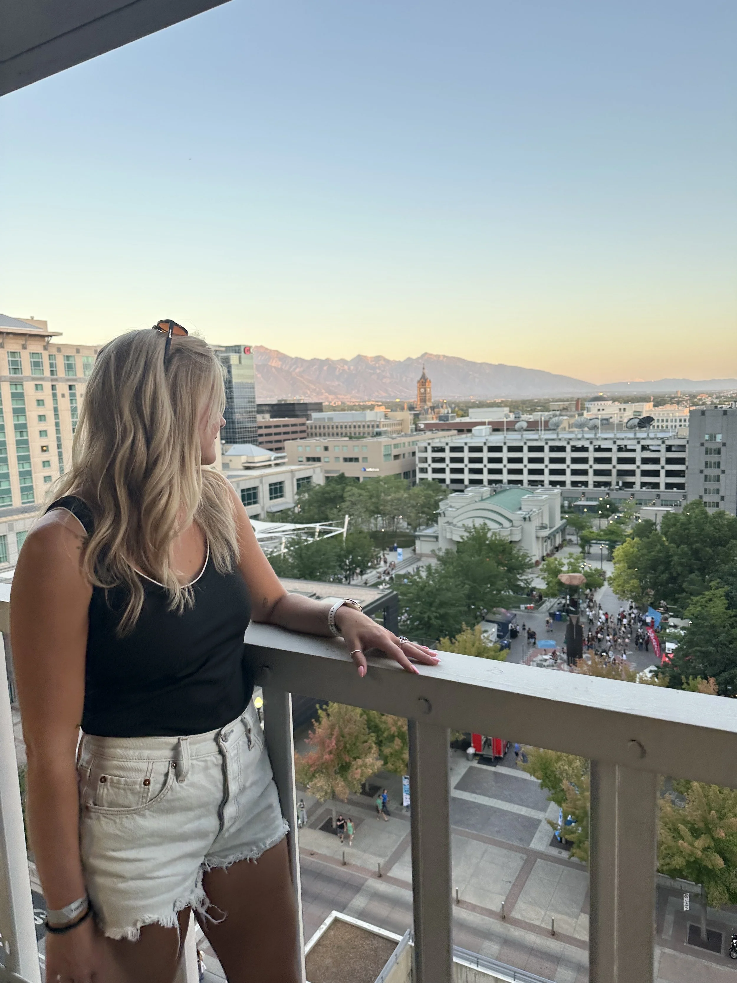 A young woman with long blonde hair wearing a black tank top and white ripped shorts, standing on a balcony, looking out over a city with buildings, a clock tower, trees, and mountains in the background during sunset.