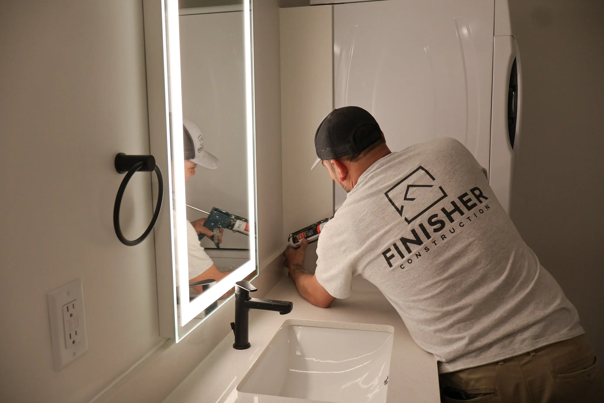 A man wearing a gray T-shirt and a black cap is installing or repairing a light fixture above a bathroom sink and mirror. The mirror is illuminated and reflecting his image, and he's using tools. The bathroom has a white countertop, a modern black faucet, and white walls with electrical outlets.