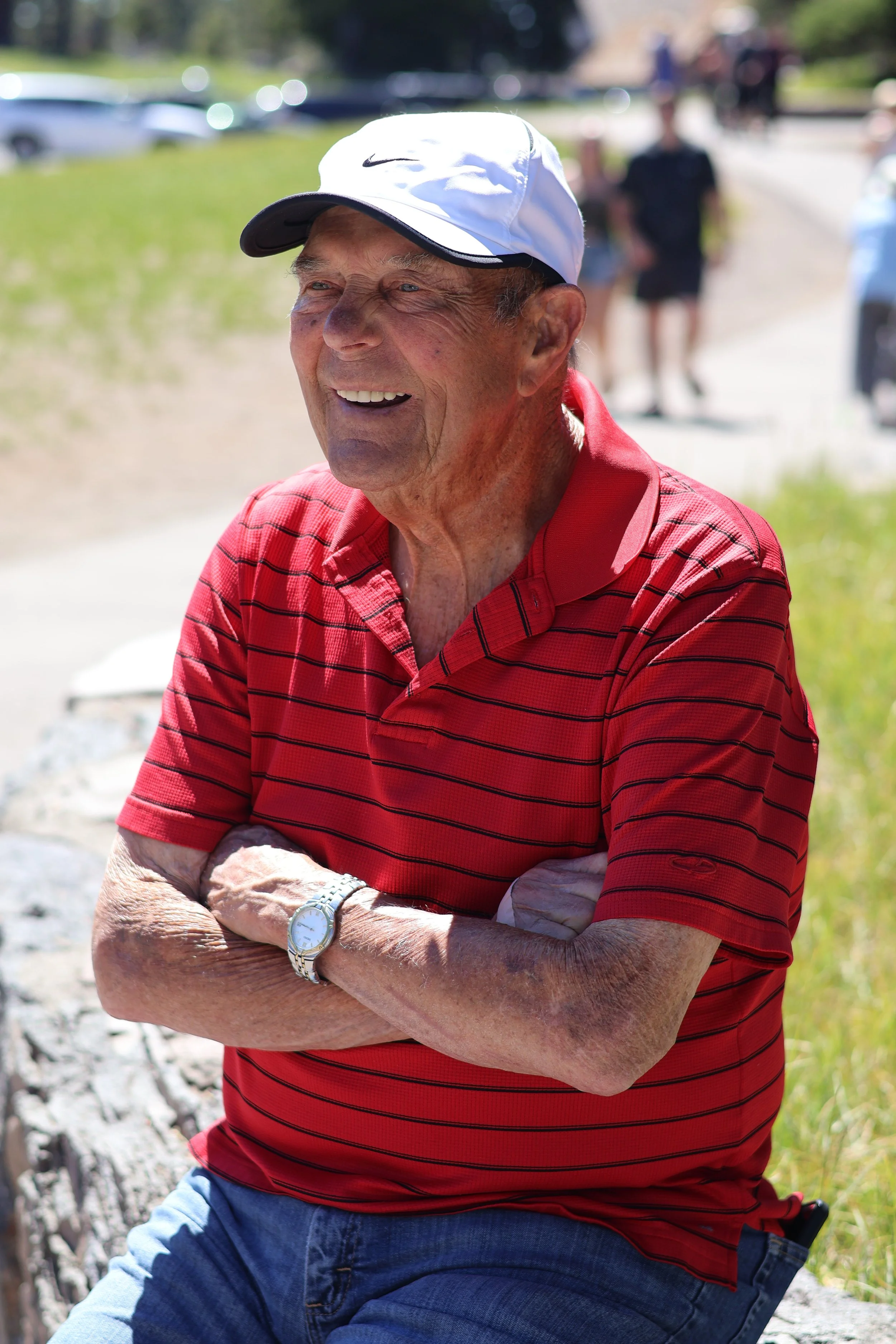 A smiling elderly man with wrinkles, wearing a white Nike cap, a red and black striped polo shirt, and blue jeans, sitting outdoors with arms crossed. There are people and cars blurred in the background on a sunny day.