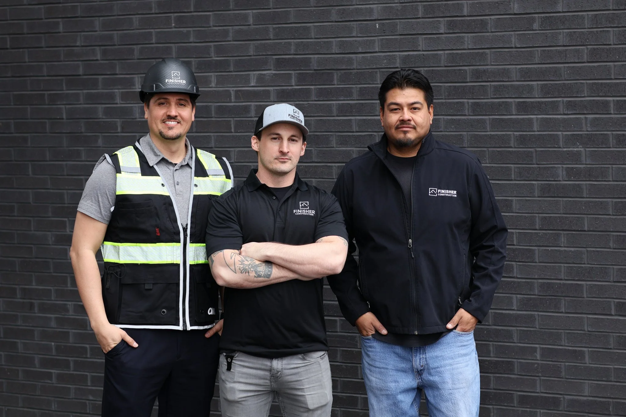 Three construction workers standing in front of a dark brick wall, all wearing work attire with the 'Finisher Construction' logo. The worker on the left is wearing a helmet and a safety vest, the one in the middle has a baseball cap and decorated tattoos, and the one on the right is wearing a jacket.