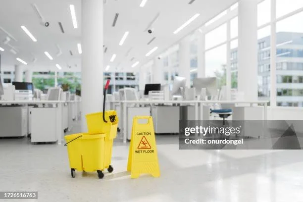 Empty office with a wet floor sign and cleaning mop in the foreground, modern desks and chairs in the background.