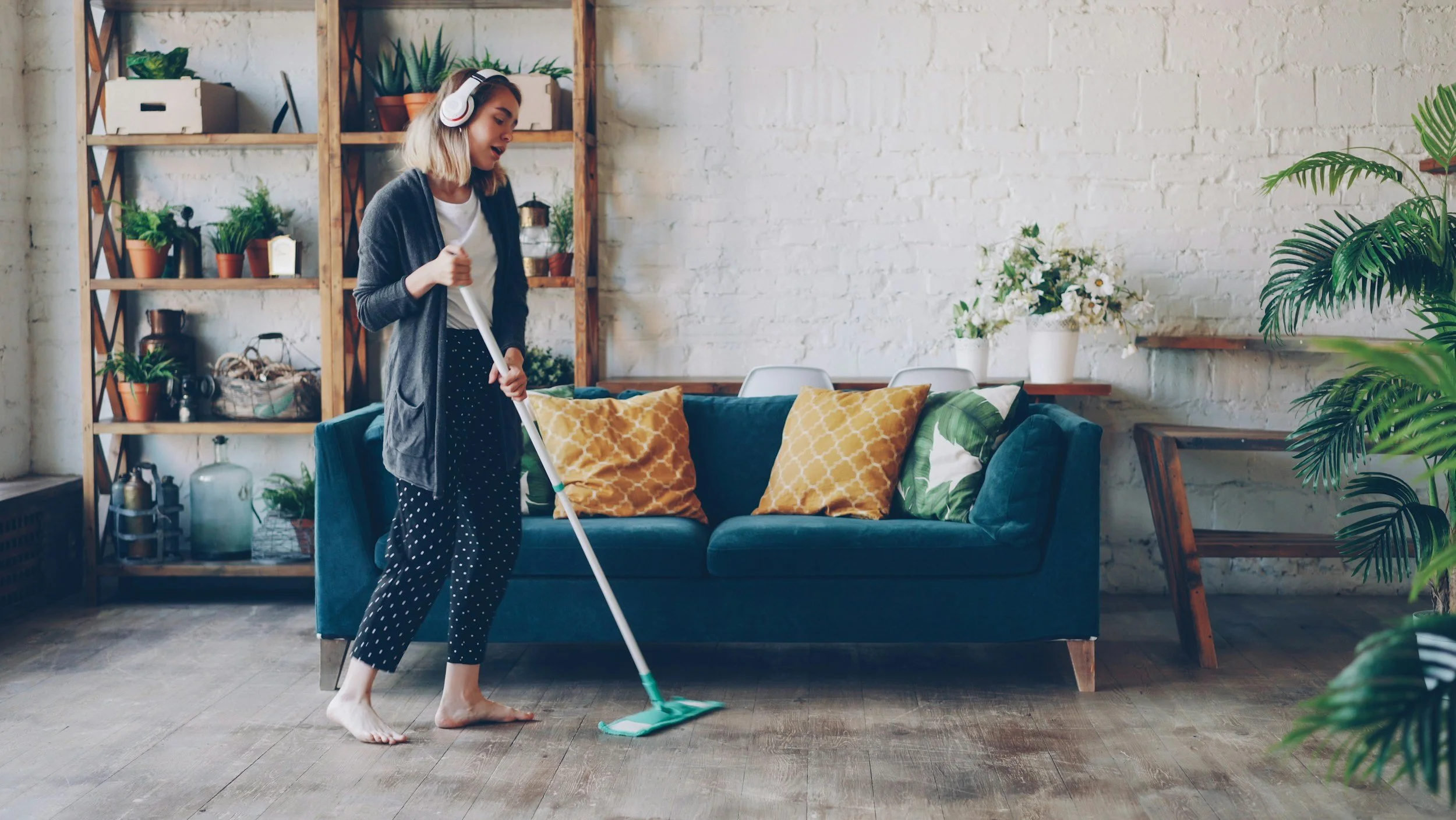A woman with headphones mopping the wooden floor of a living room with a blue sofa and yellow pillows, surrounded by potted plants and a white brick wall.