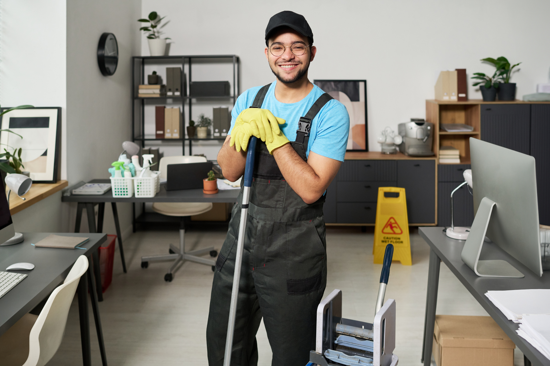 A man in cleaning uniform, yellow gloves, and a cap smiling and holding a mop in an office setting with desks, chairs, computers, and a caution sign on the floor.