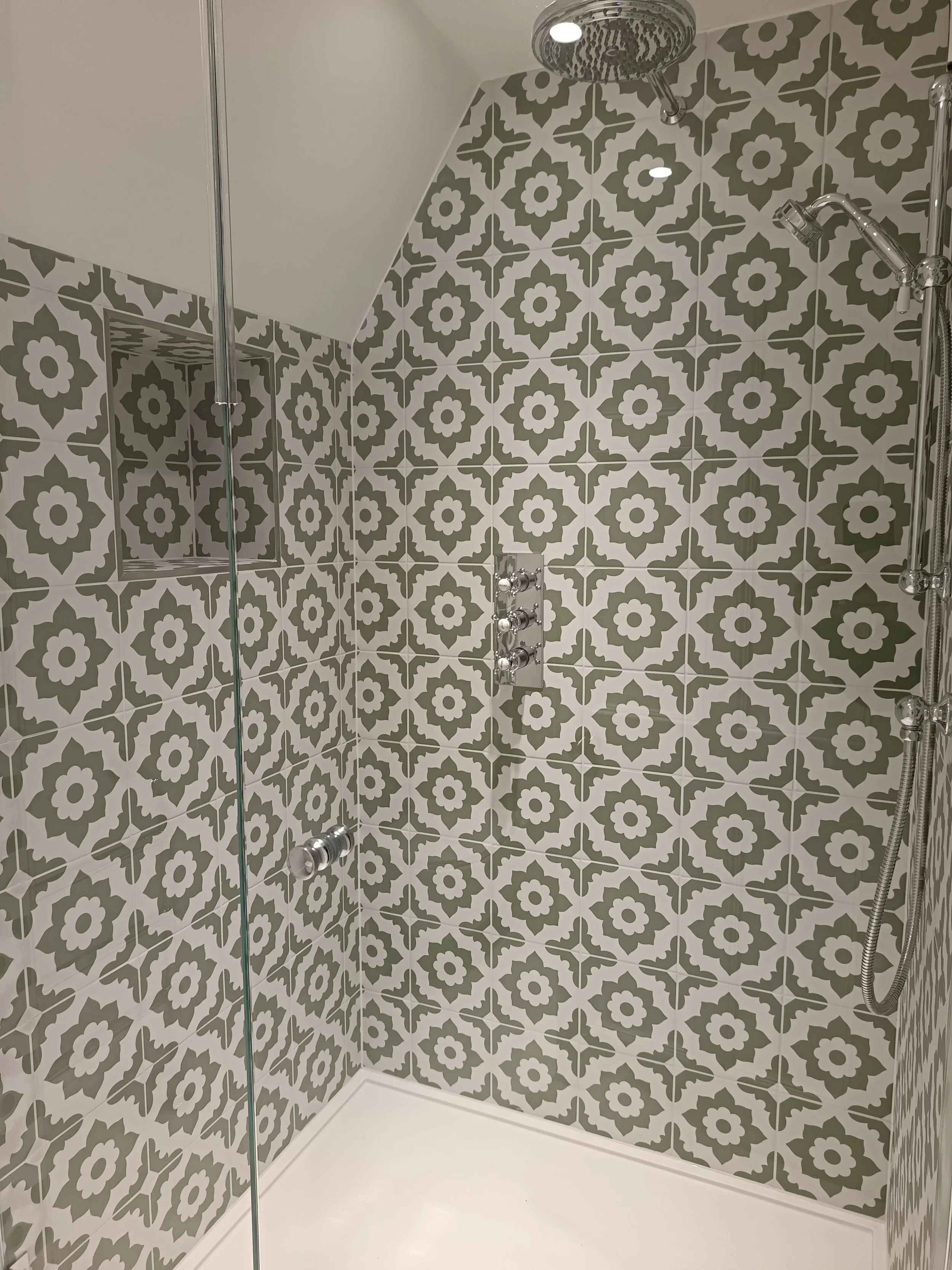 Shower area with patterned tiles on the wall, a chrome overhead shower, a wall-mounted faucet panel, and a handheld showerhead with a hose.