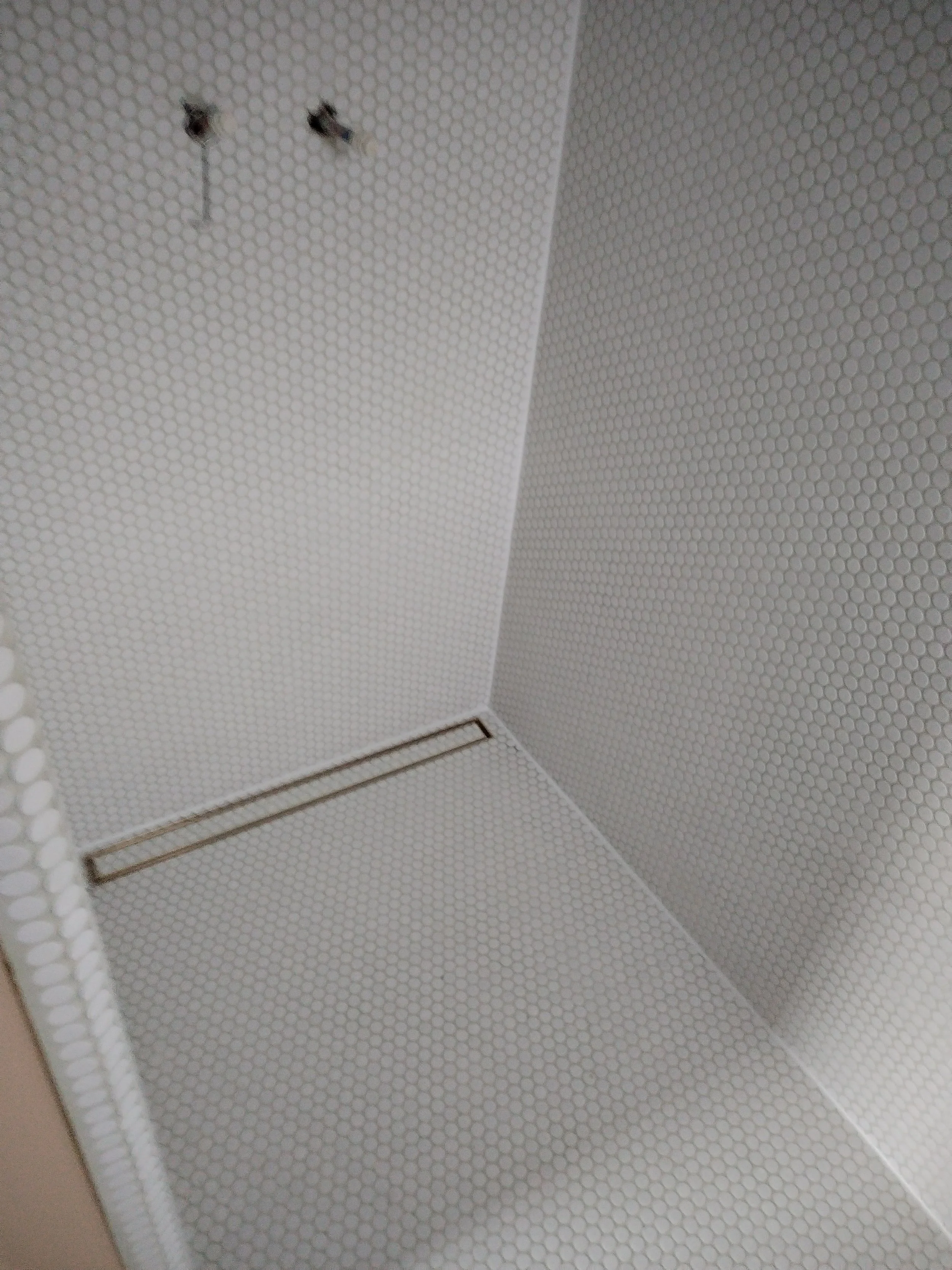 Close-up of a corner inside a room with white hexagonal tile walls and floor, featuring two holes in the wall and a rectangular drain on the floor.