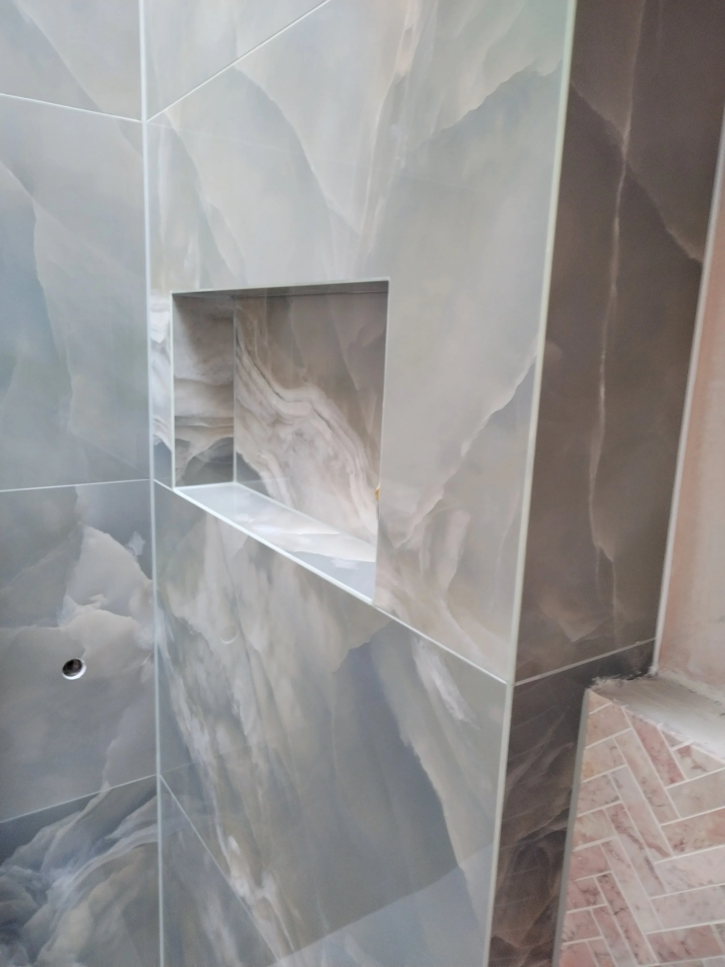 A bathroom wall with large marble tiles and a small recessed shelf near the bottom. The tiles have a grey and white marble pattern. The floor has smaller tiles with a brick-like pattern.