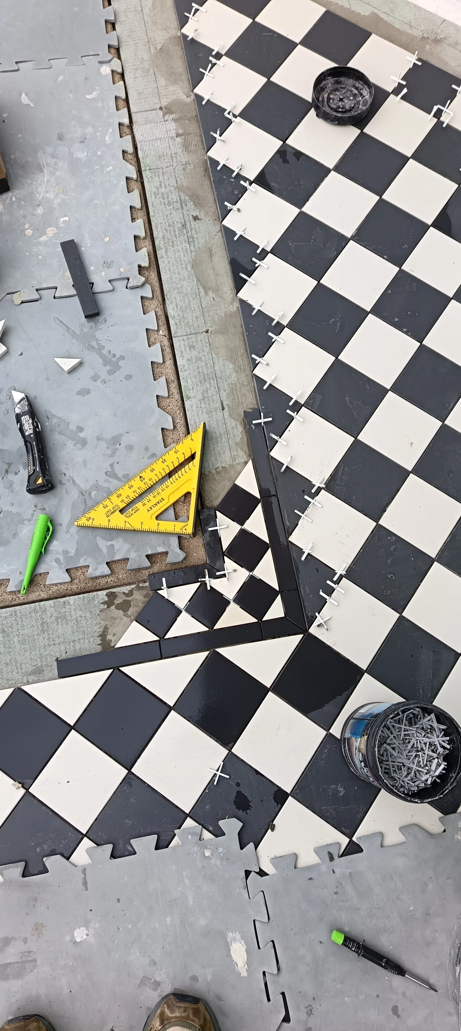 A partially completed black and white checkered tile floor being installed. Tools, including a ruler, a utility knife, and a green marker, are on the floor. Spacers are used between tiles for even spacing, and a bucket of extra spacers is visible.