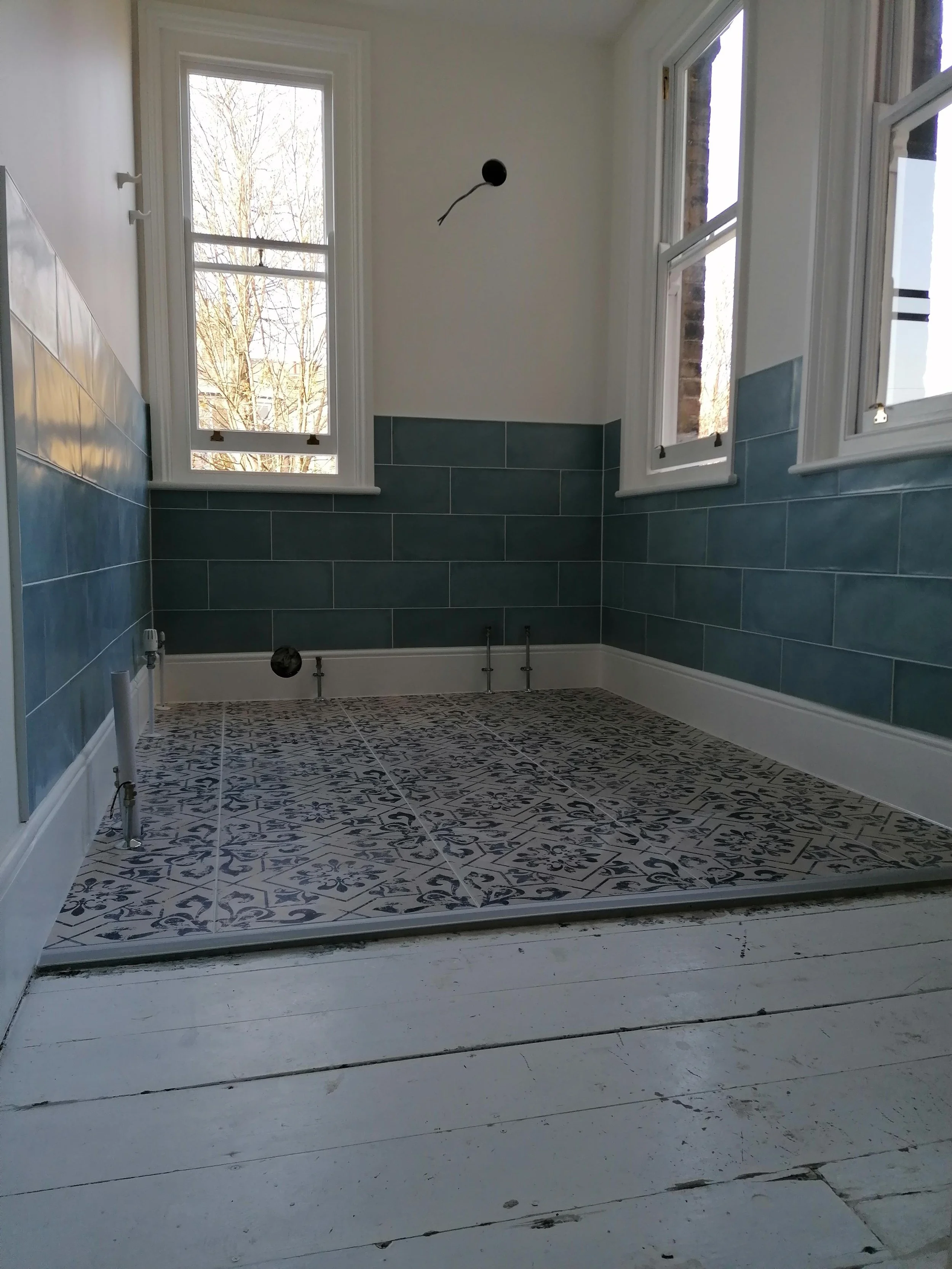 Empty room or bathroom under construction with blue tiled walls, patterned floor tiles, white windows, and exposed pipes, with a white wooden floor in front.