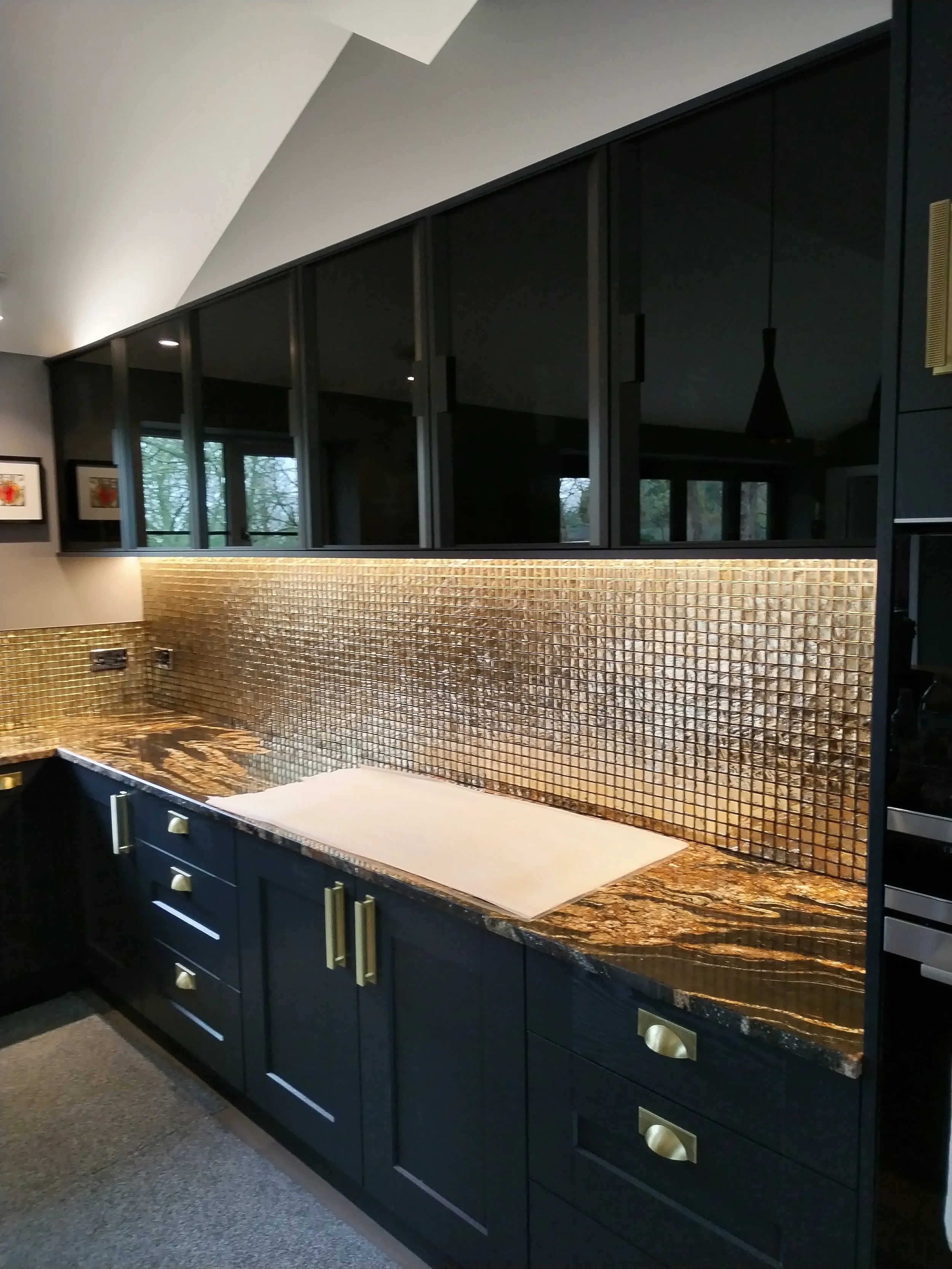 Dark blue kitchen cabinets with gold handles, a gold textured backsplash, and a granite countertop in shades of brown, black, and gold, with a white cutting board on the counter.