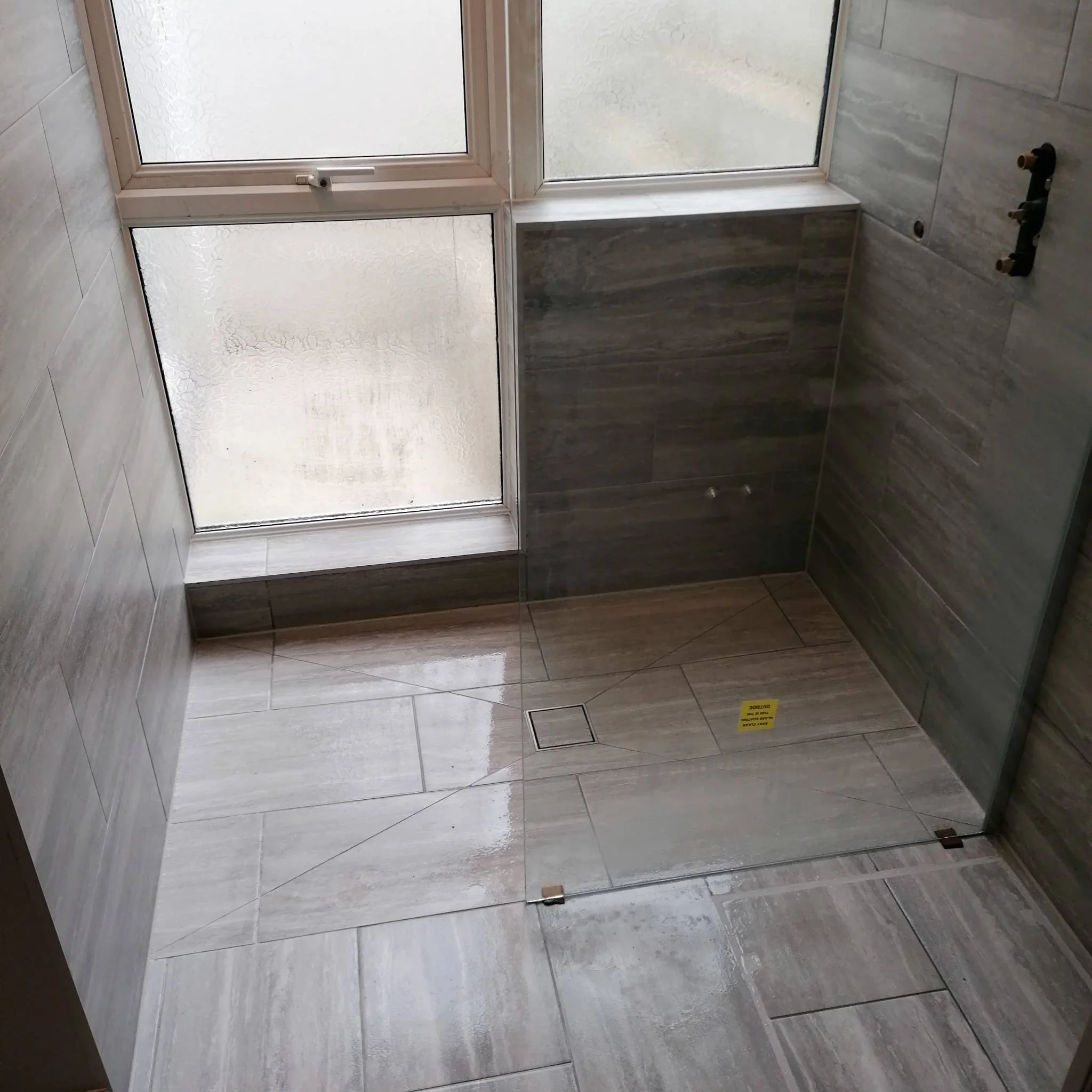 A tiled bathroom with frosted glass window on the upper wall, a small window ledge below, and a drain cover on the floor. There is a black fixture on the right wall, possibly for plumbing.