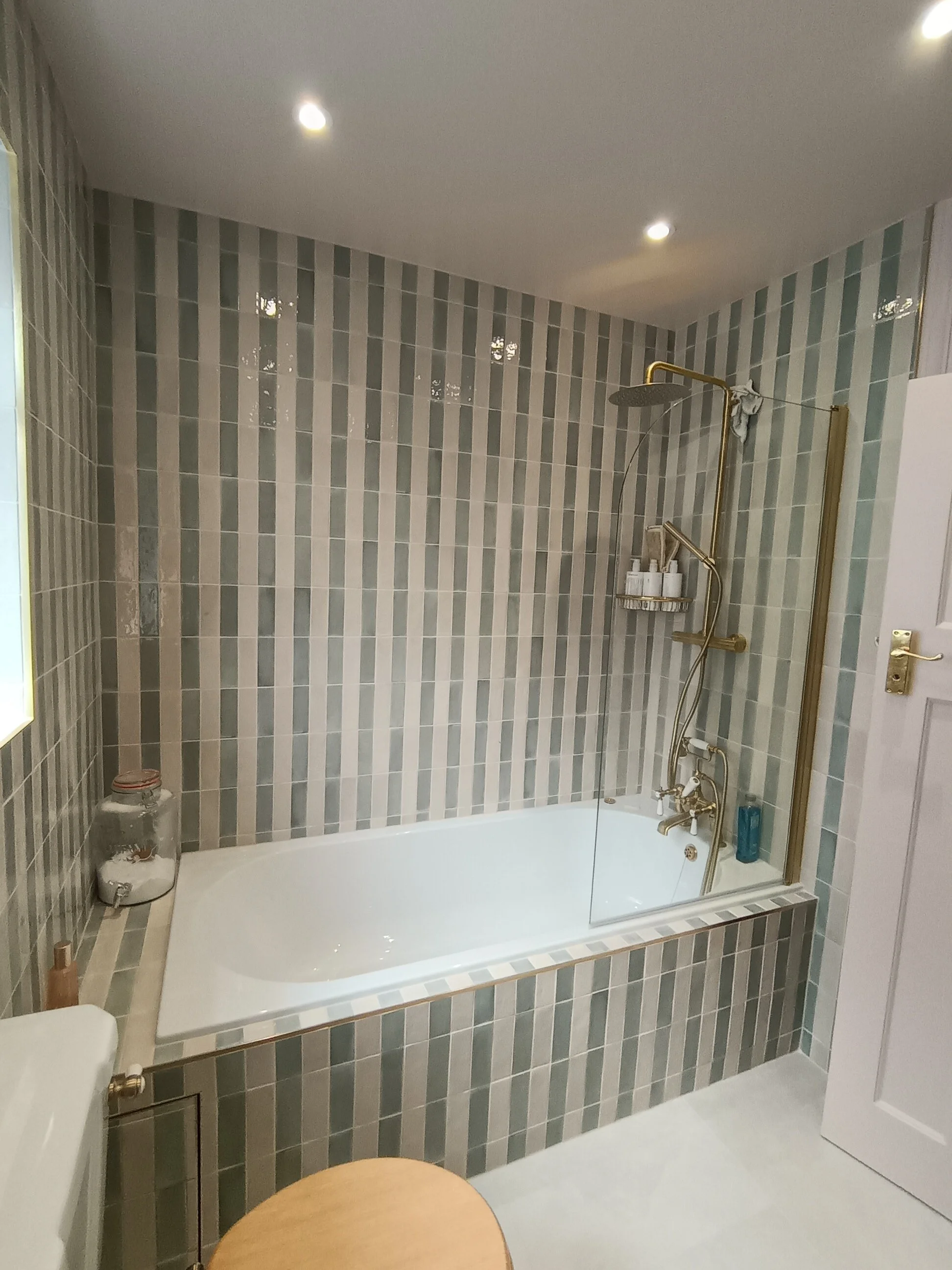 Bathroom with bathtub, gray and white striped tiles on the wall, overhead lighting, gold shower and faucet fixtures, small shelf holding toiletries, glass shower door, a round wooden table in the foreground, and a large jar with towels.