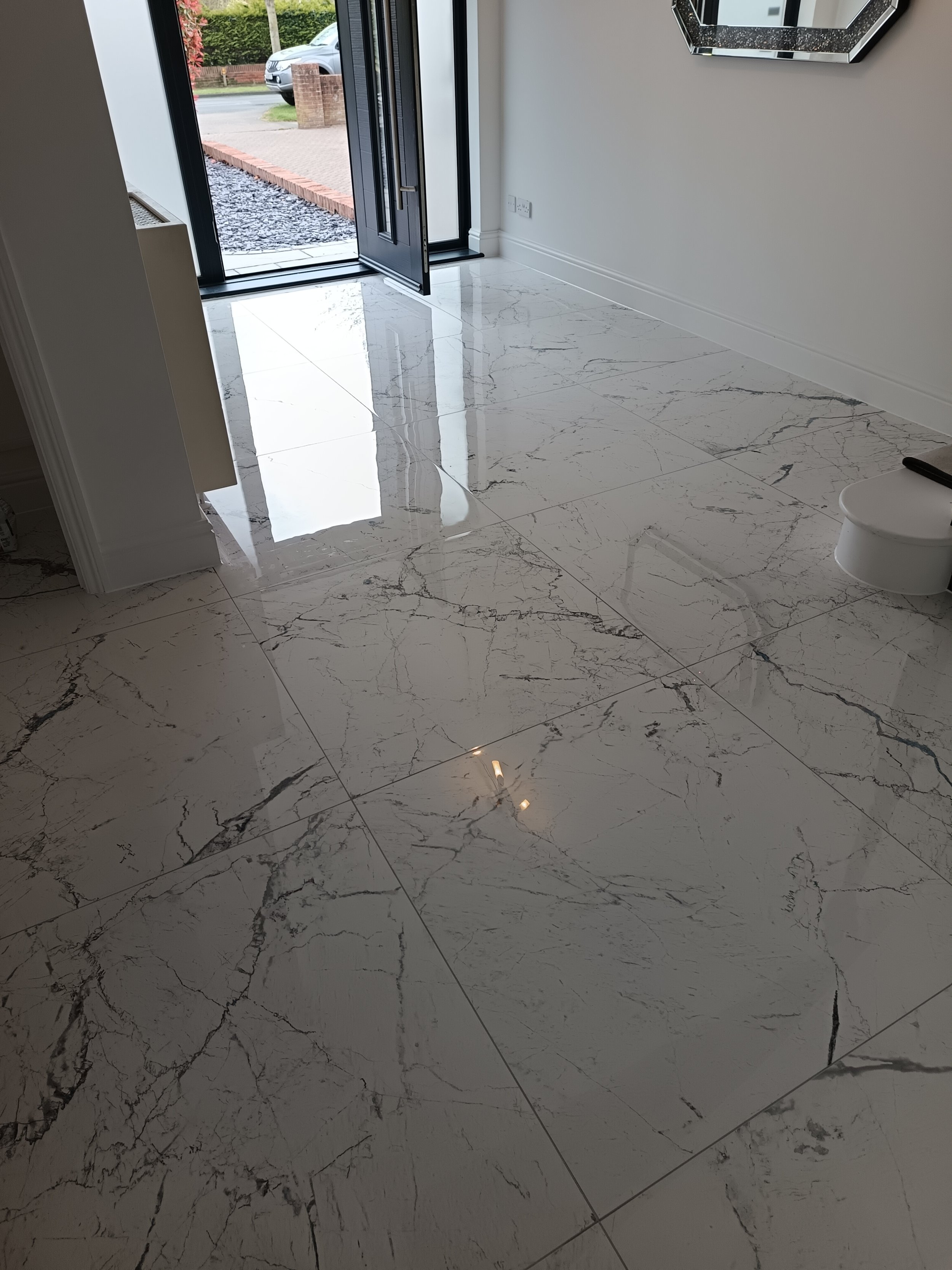 A modern interior space with white marble flooring featuring dark veining, a black front door with glass panels, and a white wall with a decorative mirror.