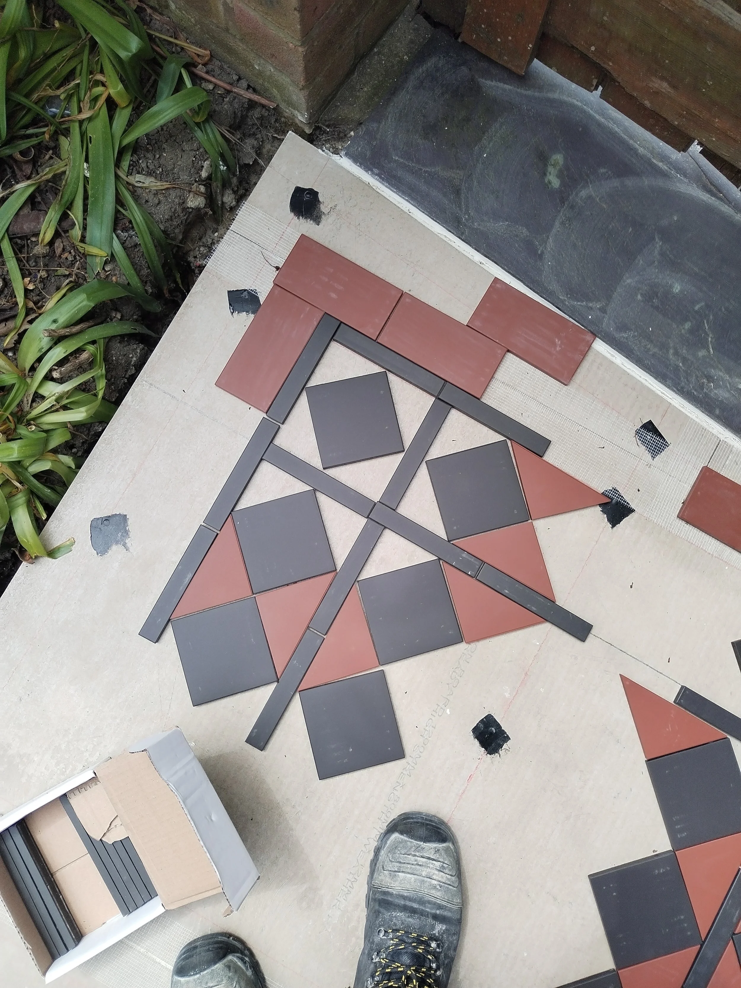 A person standing on a tiled floor wearing black work boots with yellow laces. There are some partially completed reddish-brown and gray tile pieces arranged on the floor, likely for a tiling project. Some tiles are stacked in a box to the left, and nearby there is a garden with green plants and soil, along with a brick wall.