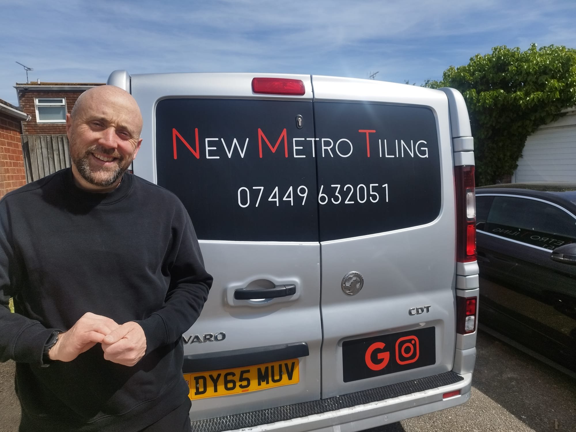 A man smiling and standing in front of a silver van with the words "New Metro Tiling" and a phone number on the back door.
