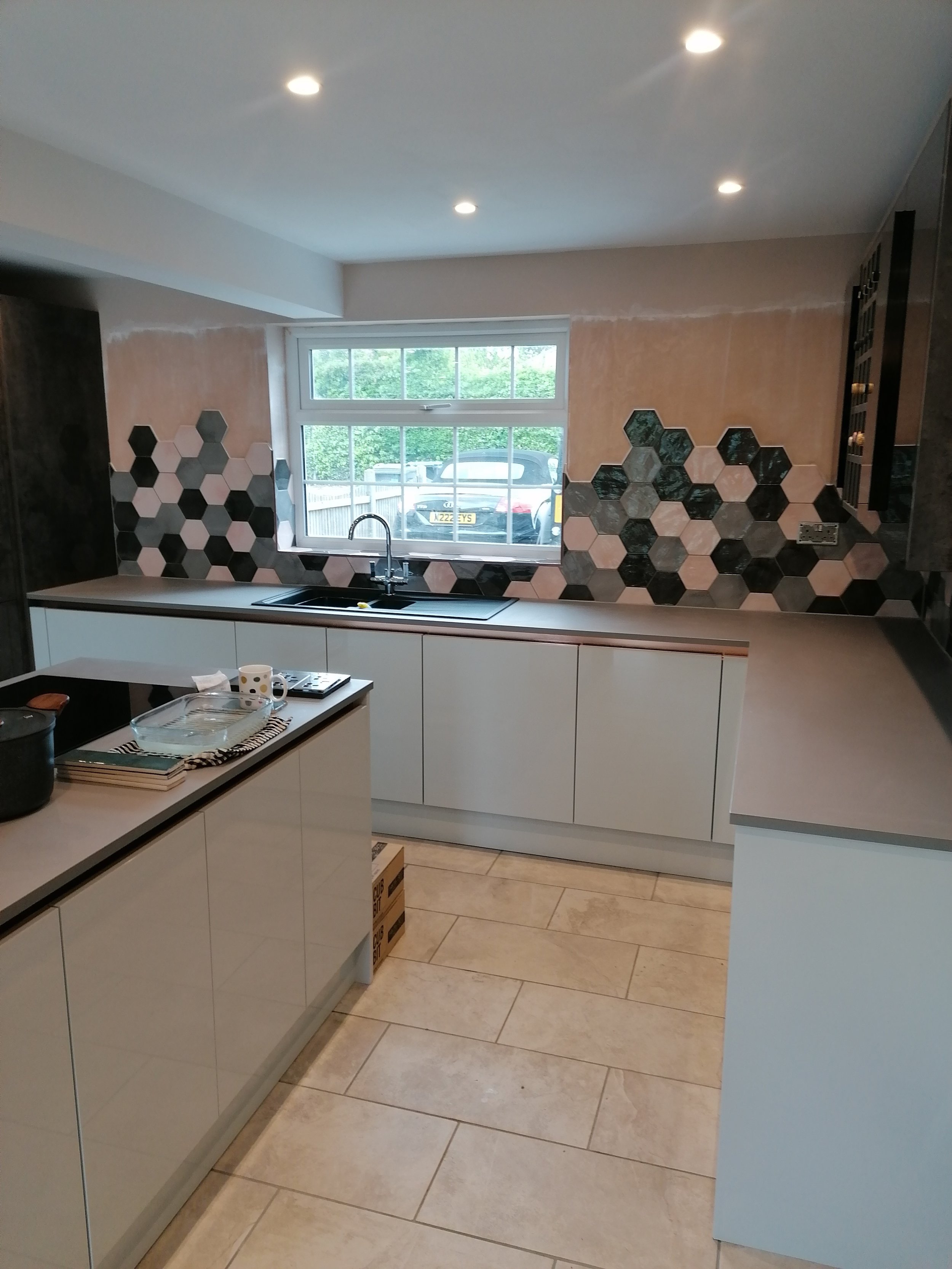 New kitchen with light-colored cabinets, hexagon tile backsplash in shades of gray, black, pink, and beige, and a window above the sink overlooking a driveway with parked cars. The ceiling has recessed lighting and the floor is tile.