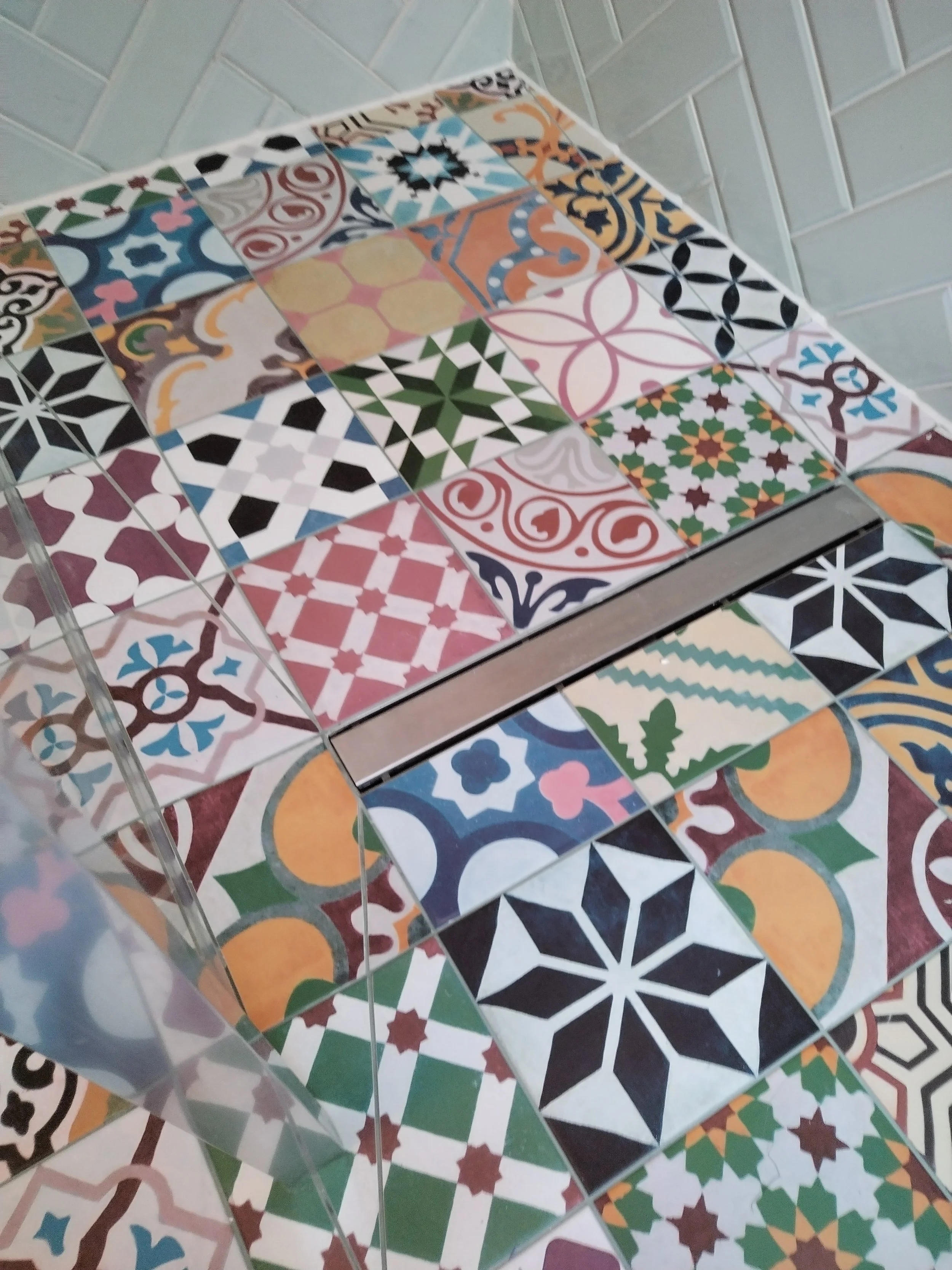 Colorful ceramic tiles with various geometric and floral patterns arranged in a grid, partially reflected on a glass surface.