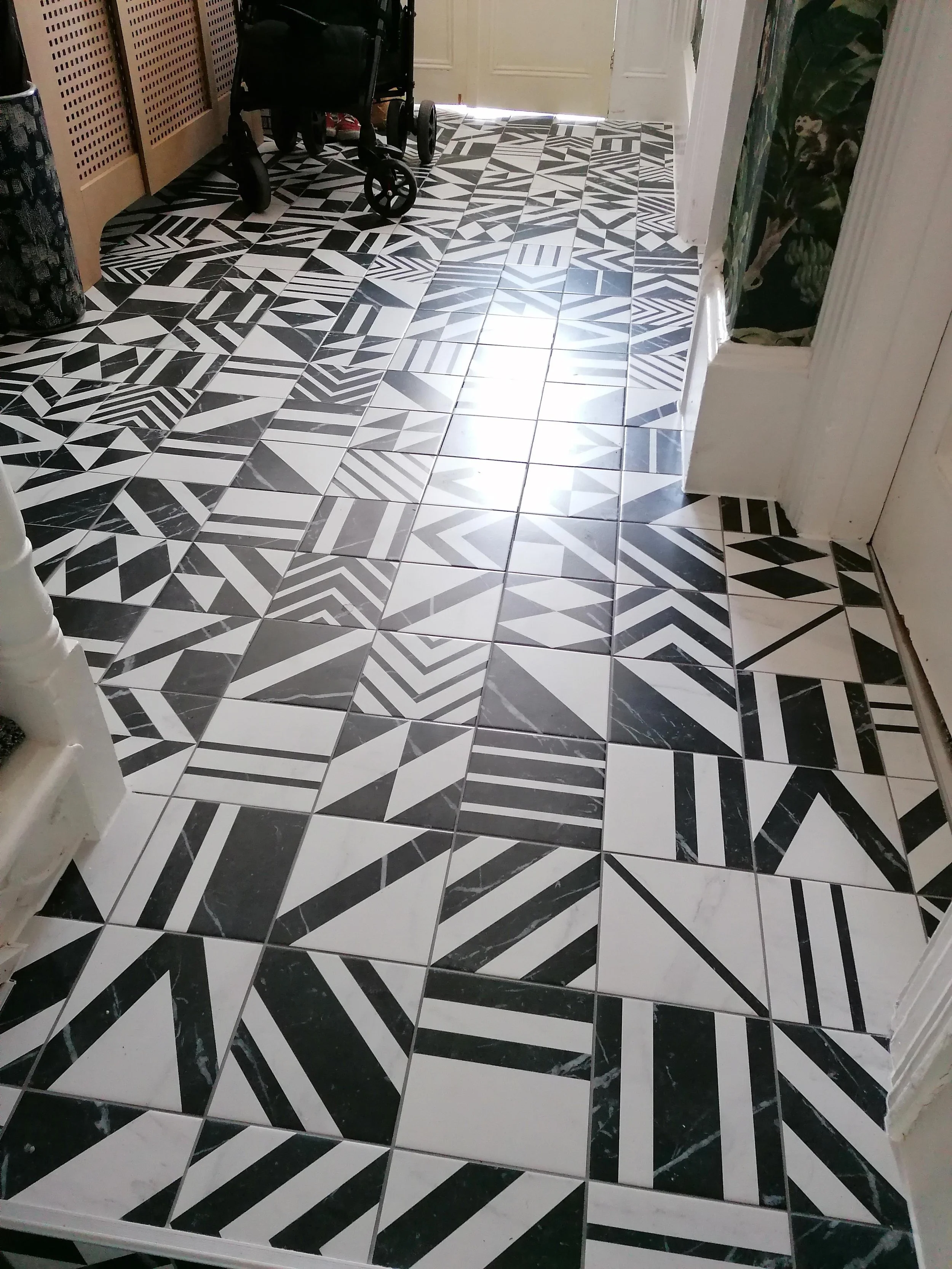 A black and white patterned tiled floor leading to a door. On the left, there is furniture with a black laundry basket on top. In the background, there is a black stroller. On the right, a wall with a floral wallpaper pattern is visible.