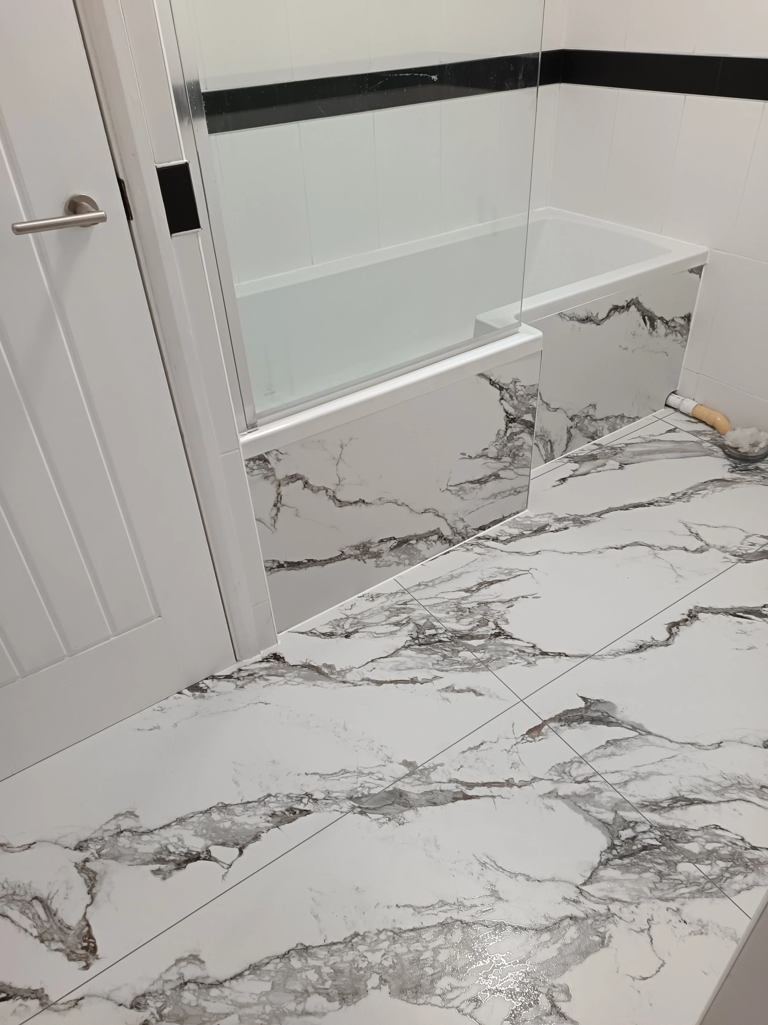 Bathroom with white marble tiles on the floor and lower wall, a partial view of a white door, a glass shower enclosure with a black and white tiled wall, and a drain on the floor.