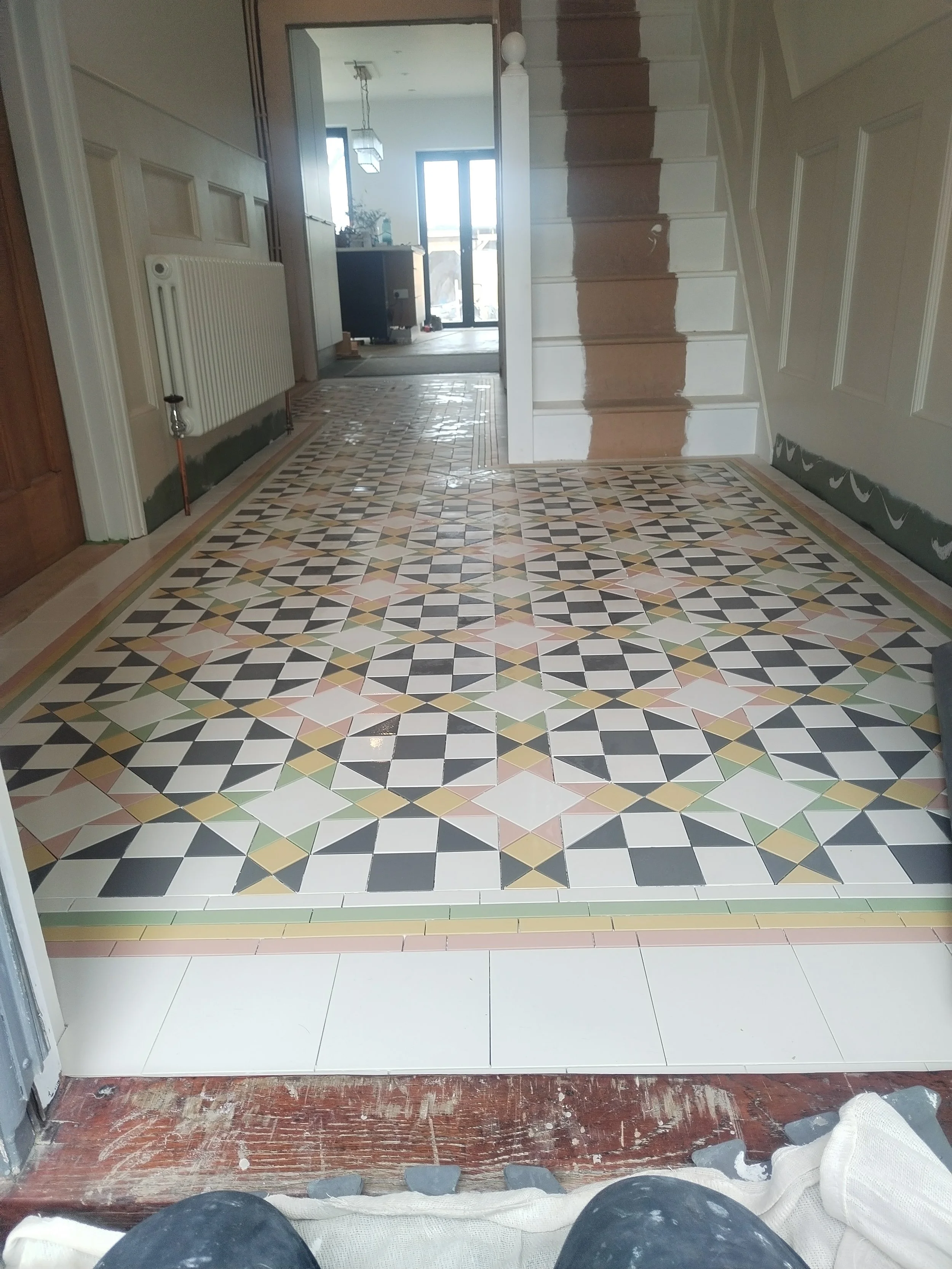 View from a person's point of view, showing a partially finished entranceway with decorative patterned floor tiles, tiled step, and a doorway leading inside a house. Construction materials and tools are visible.