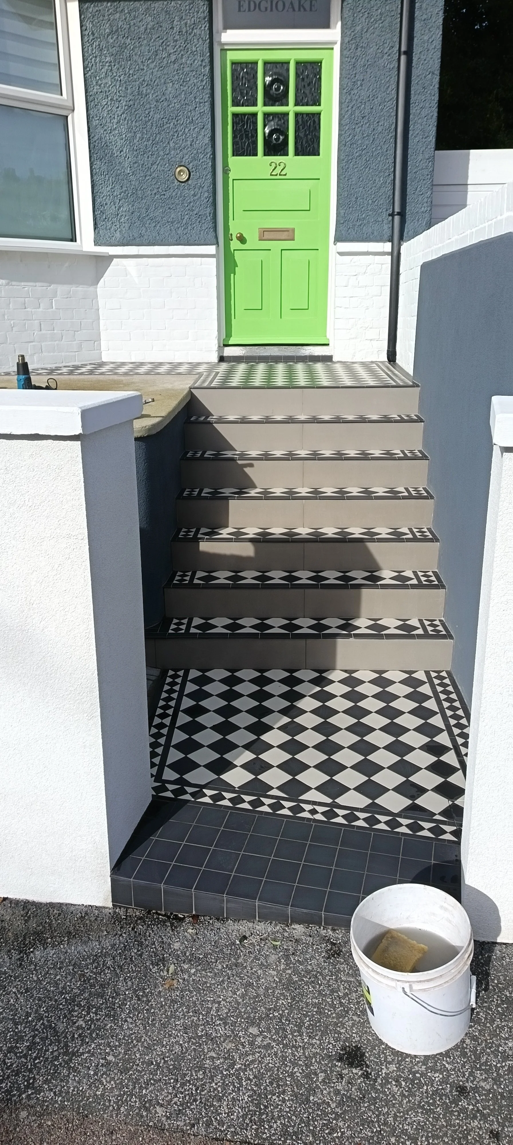 Front porch of a house with black and white patterned tiled steps leading to a bright green front door. The steps and landing have matching tiles, and there is a white wall on either side of the stairs. A bucket with a sponge is on the gravel ground 