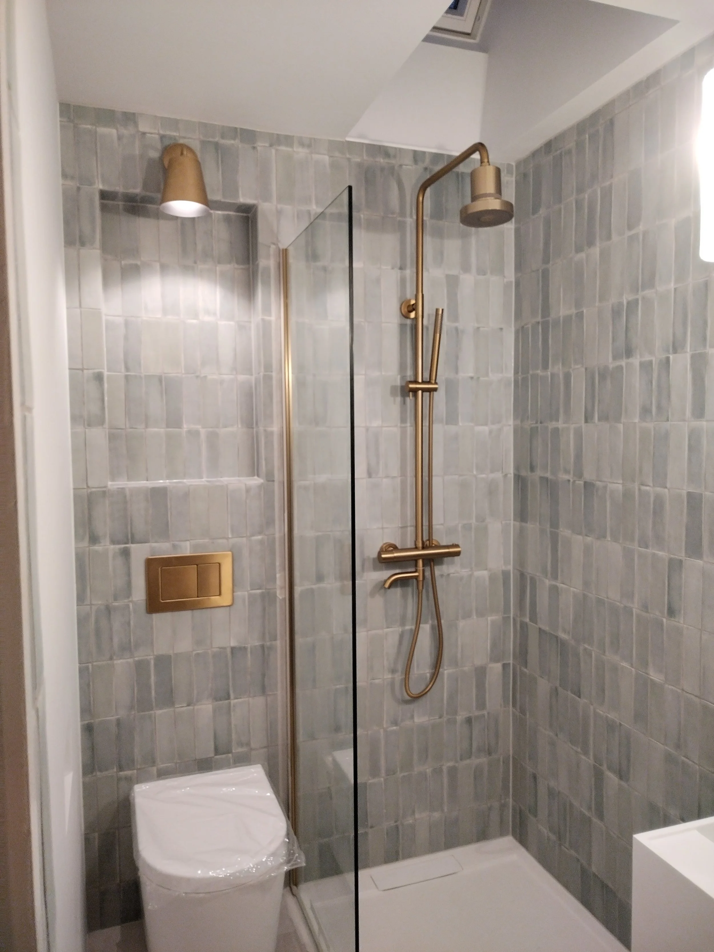 Modern bathroom with a walk-in shower featuring a gold showerhead and fixtures, gray tiled walls, a glass partition, a toilet wrapped in plastic, and a small white shelf in the corner.