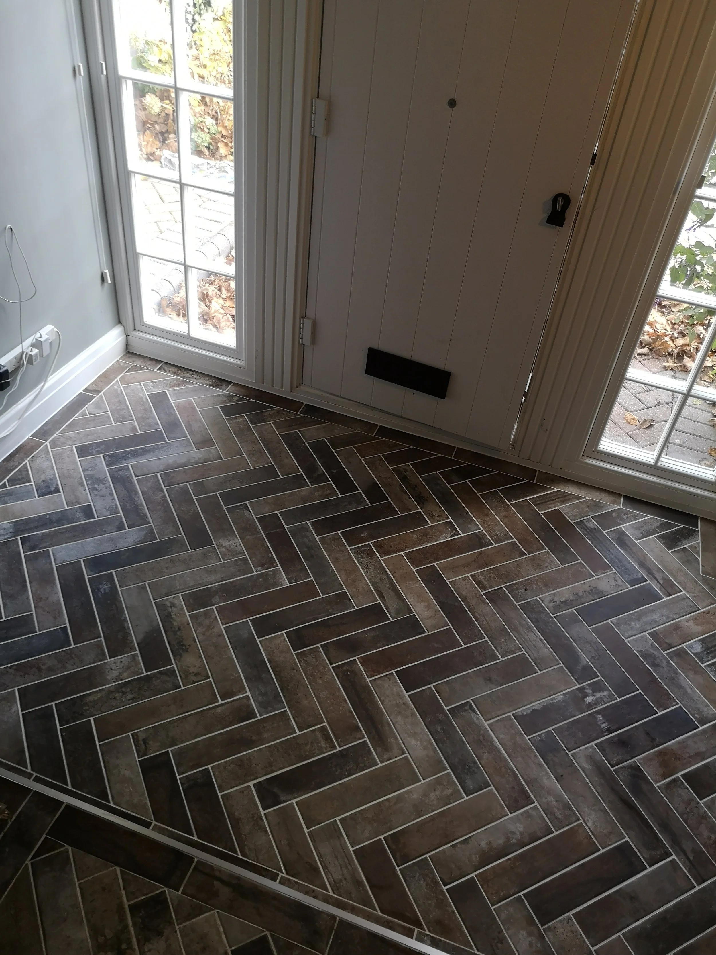 Interior of a room with a glass door leading outside, patterned tile flooring, and a white door with a black mail slot.