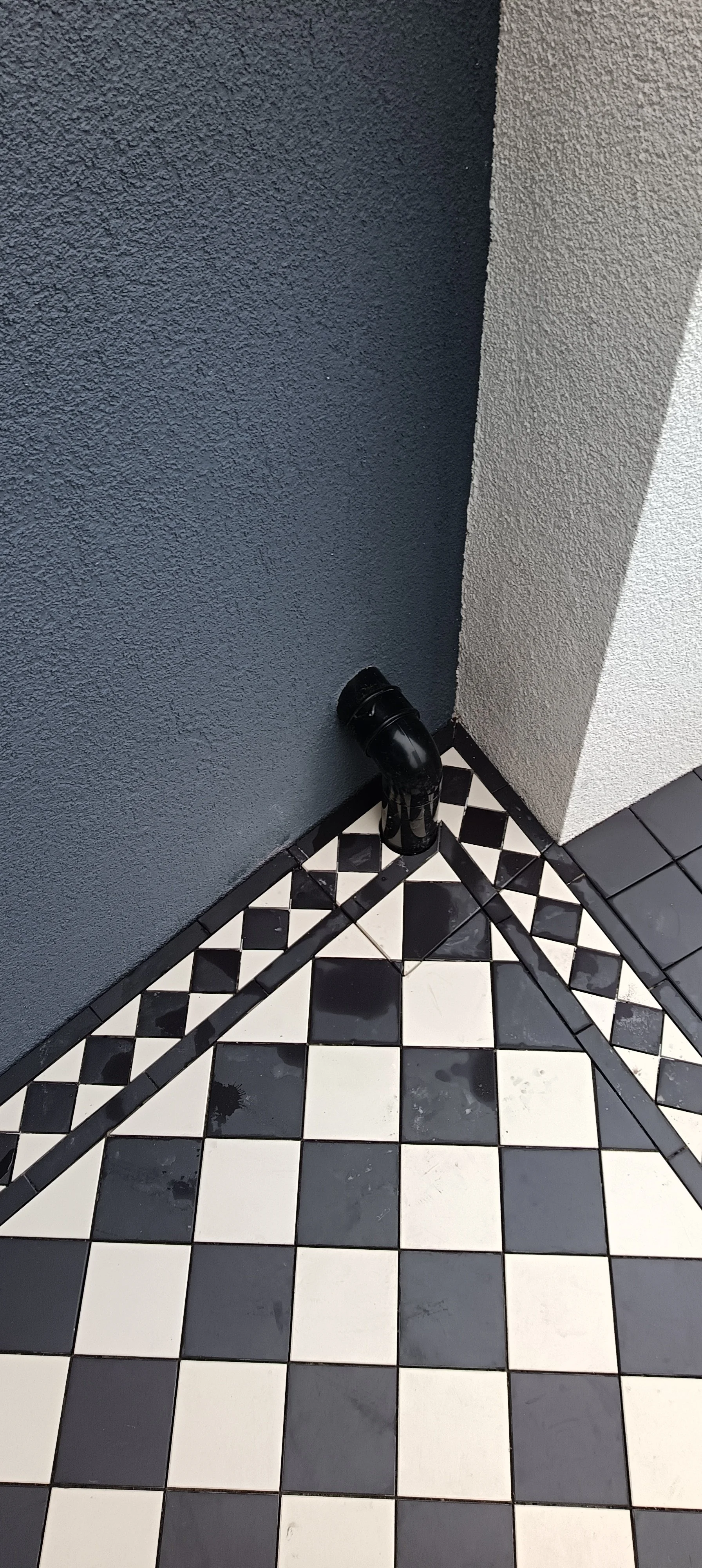 Black pipe fitting in a corner with black and white checkered tile floor and textured grey walls.