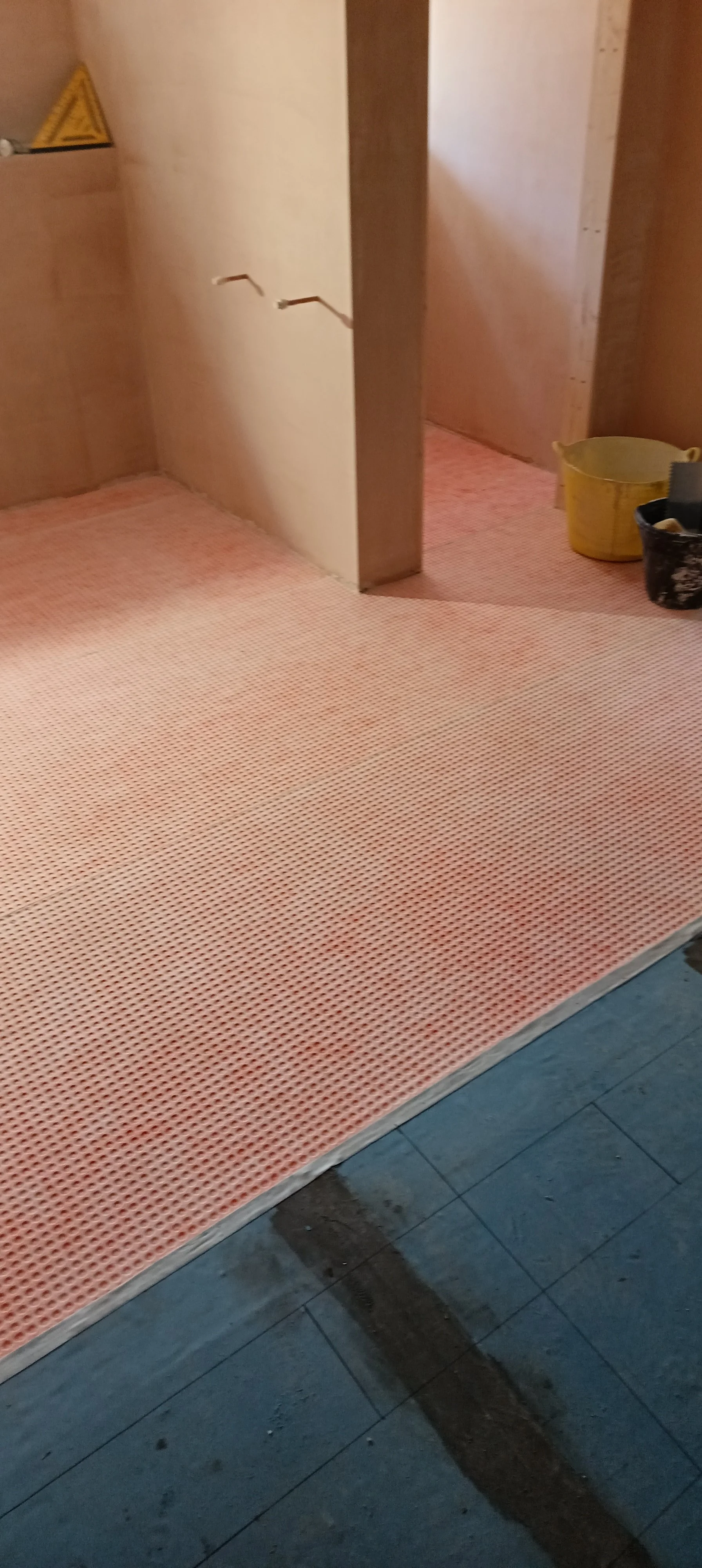 Interior construction site with pink perforated flooring, yellow and black buckets, and unfinished wooden walls.