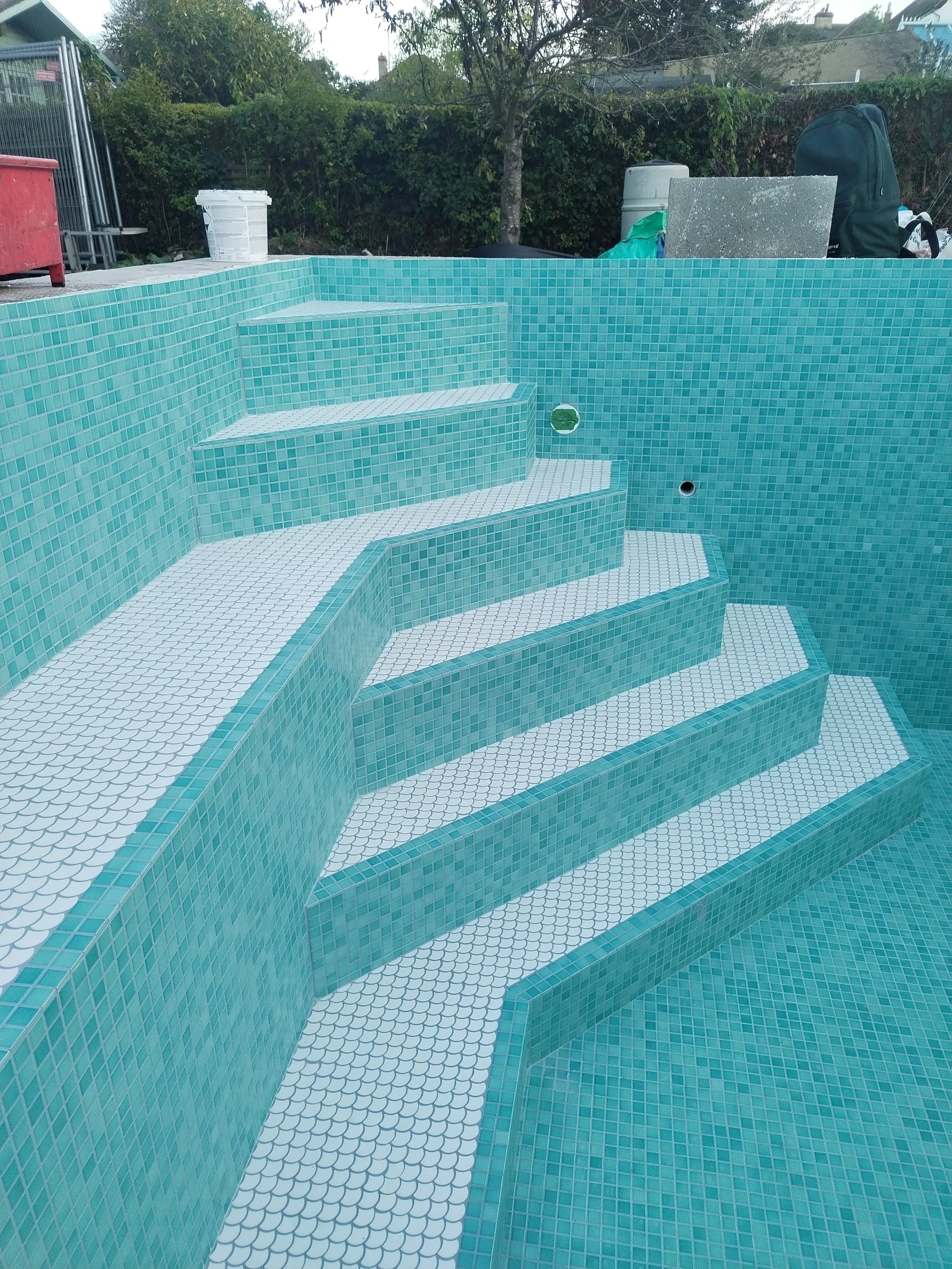 An empty swimming pool with turquoise and white mosaic tiles, featuring steps and plumbing holes, surrounded by backyard elements including a tree, bushes, and various outdoor items.