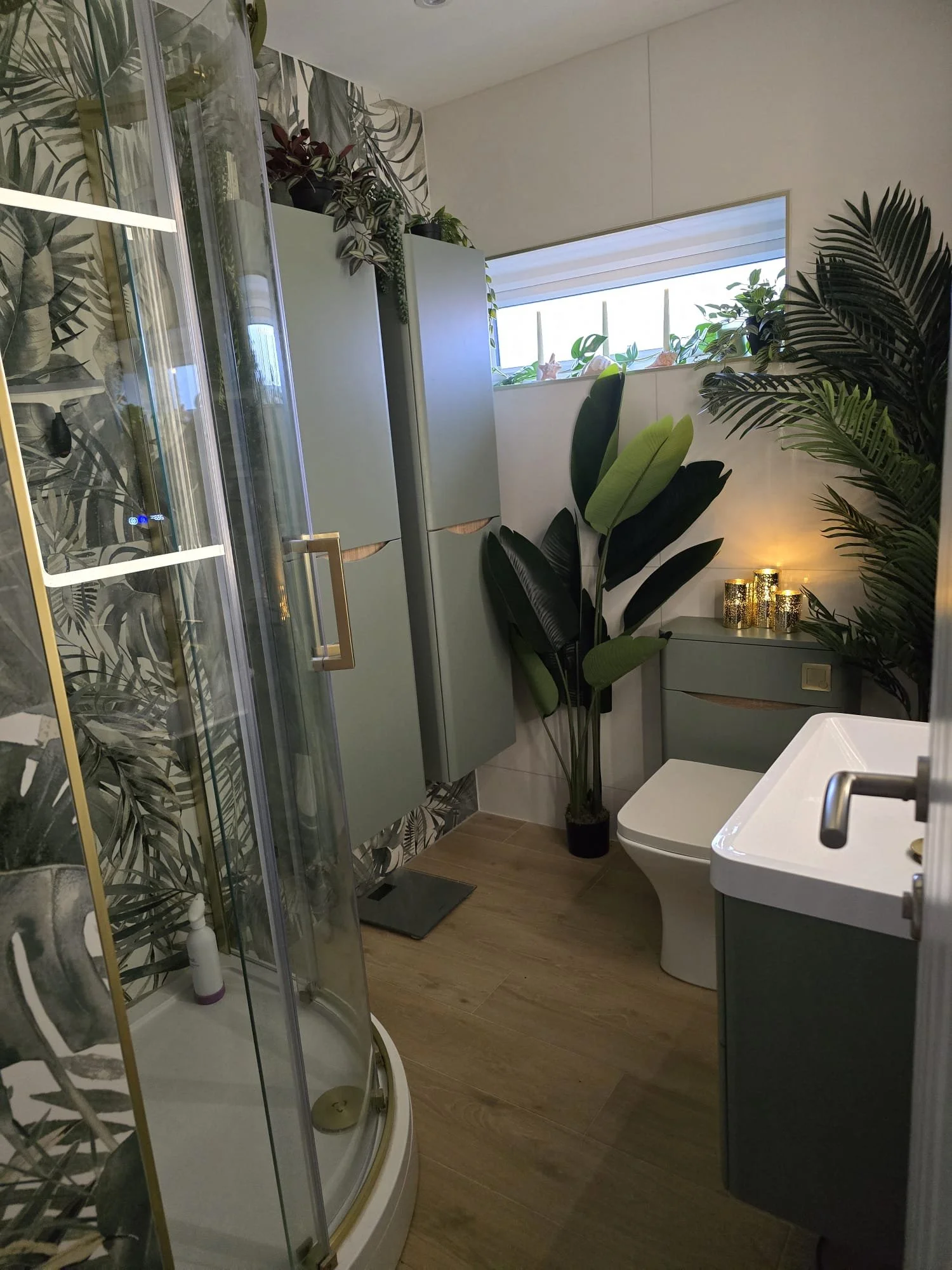 A bathroom with a shower enclosure, various plants, a window with blinds, a bidet, a bathroom cabinet, candles, and a sink.