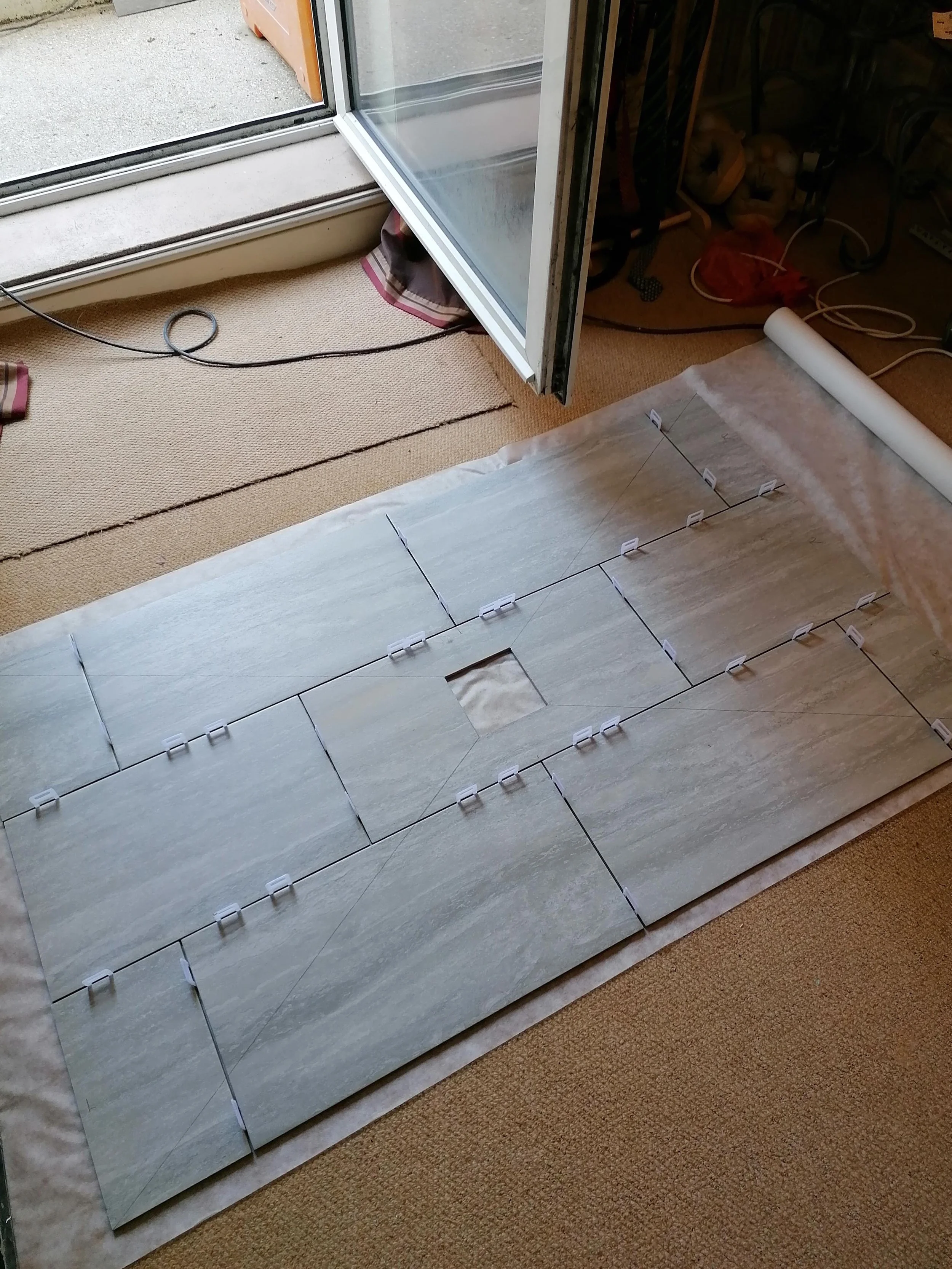 A room with beige carpet flooring and a mirror on the floor showing tiles and part of a wall. The mirror is leaning against a wall, with a roll of protective film or paper beside it. A sliding glass door with a screen leads outside, and some electric