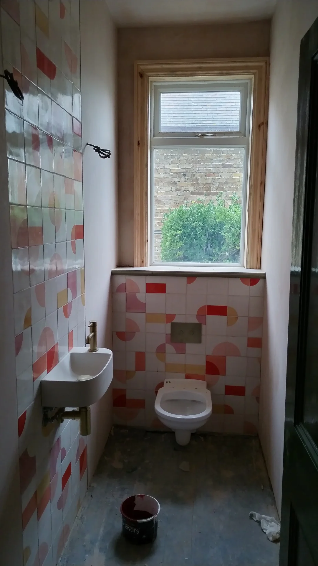 A small bathroom under renovation with a high window, patterned tiles on the wall, a wall-mounted sink, a toilet, and a bucket on the floor.