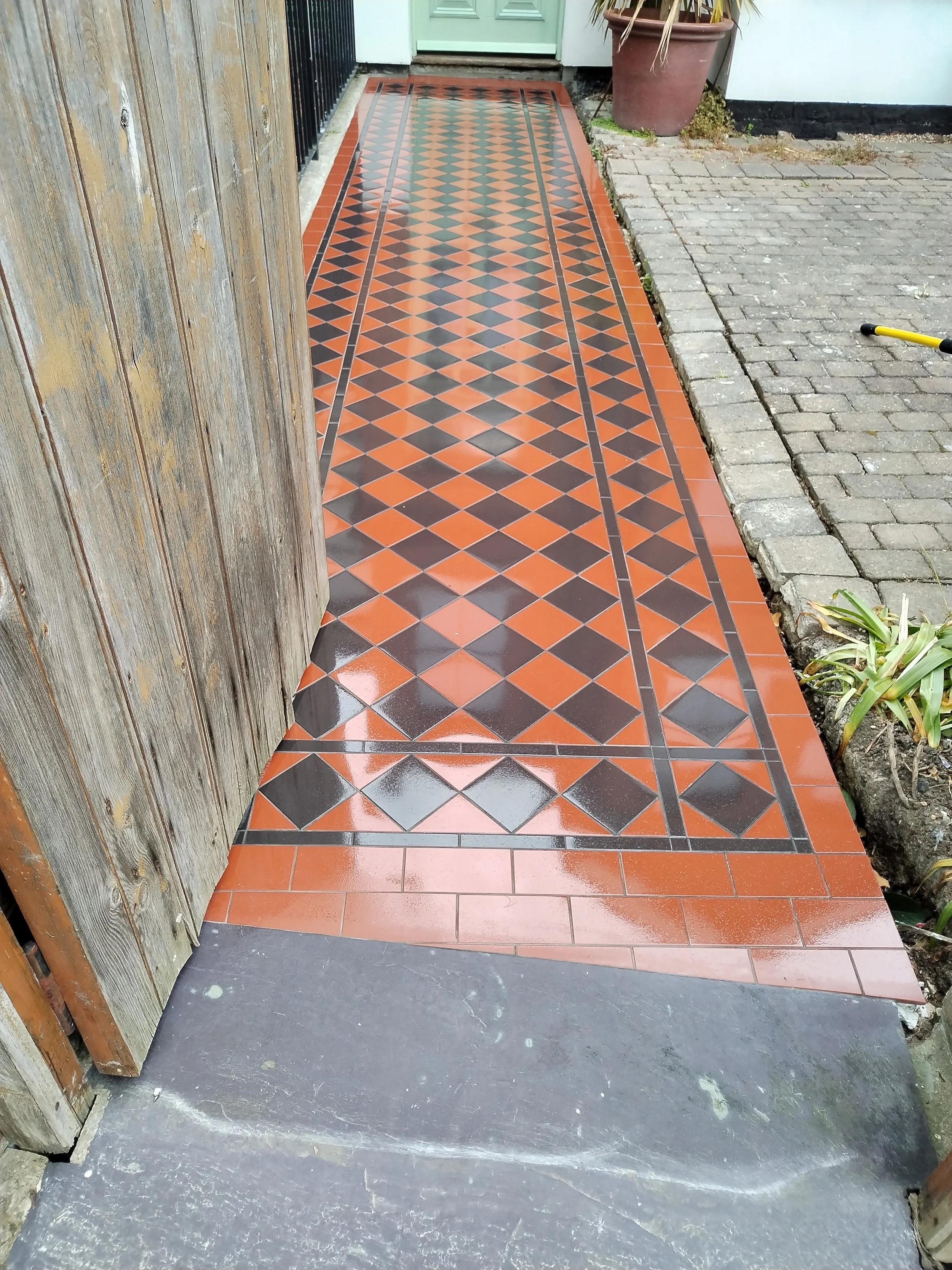 A decorative tiled pathway leading to a green door, bordered by a wooden fence on the left and a white wall on the right. There is a potted plant and some greenery near the wall.