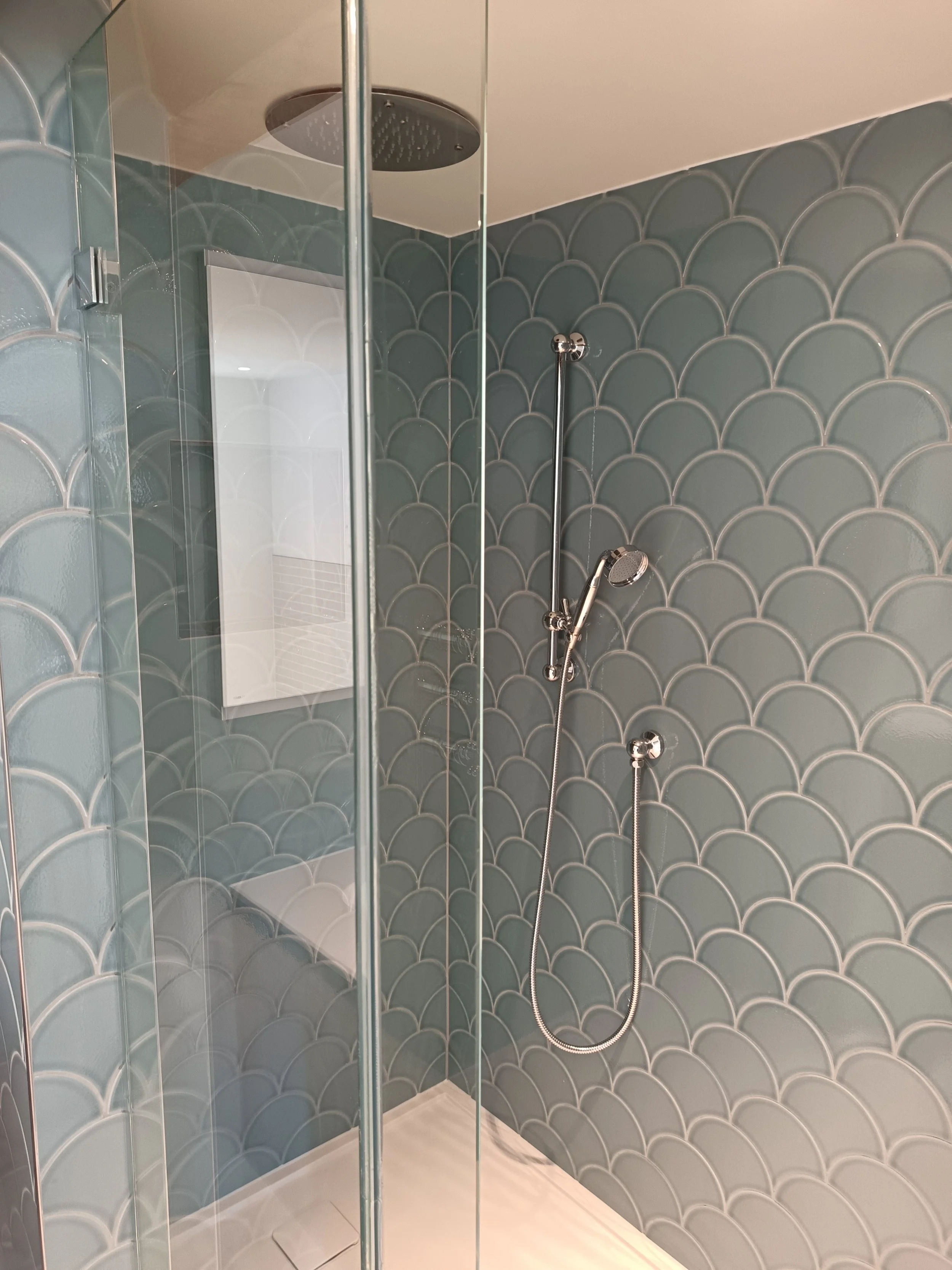 Modern shower with blue-green fish scale tiles, a rainfall showerhead in the ceiling, and a handheld showerhead on a vertical bar.