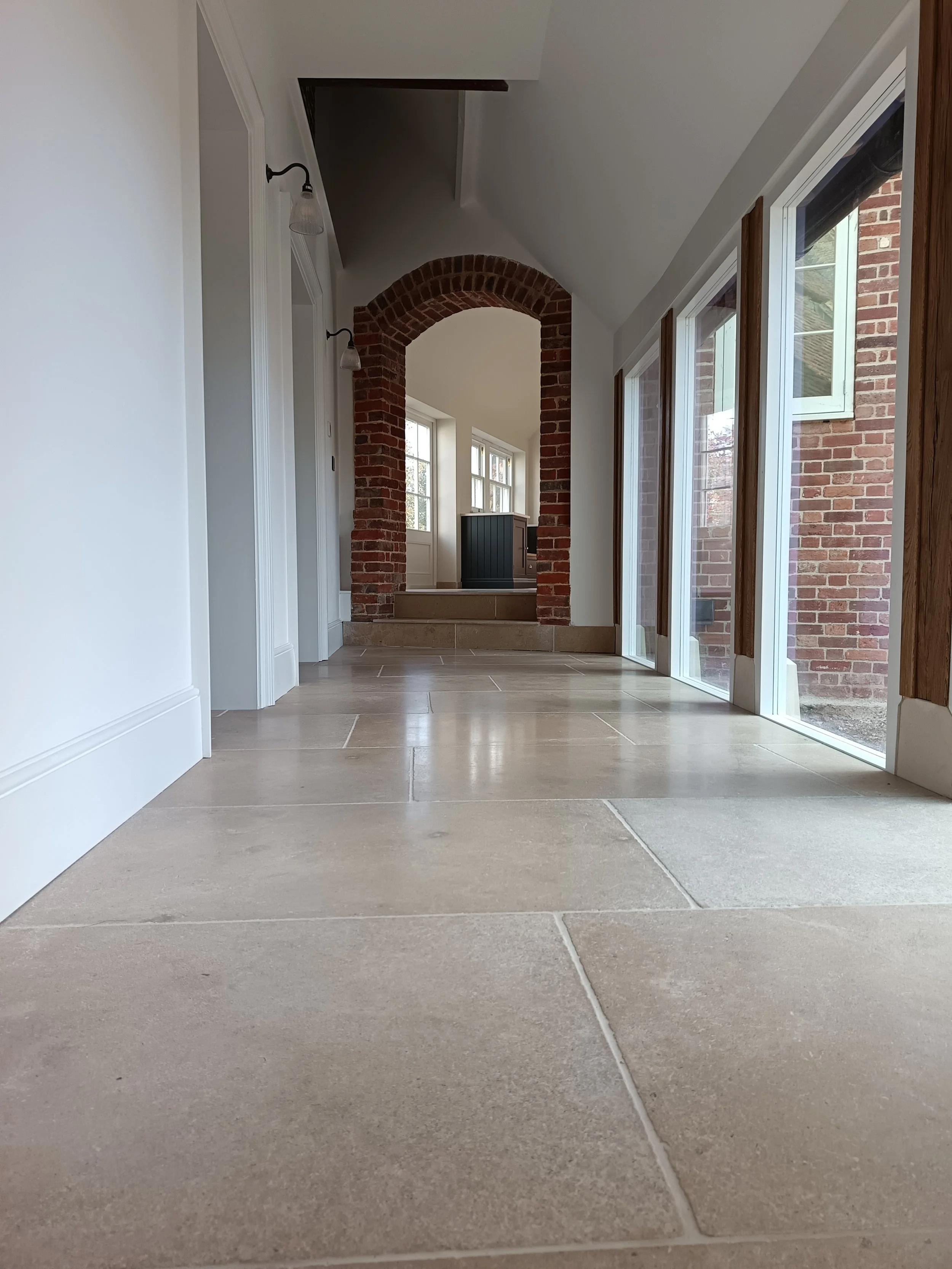 Interior view of a hallway with beige tiled floor, white walls, a brick archway, and large glass sliding doors leading outside. There are windows and wooden trim in the background.