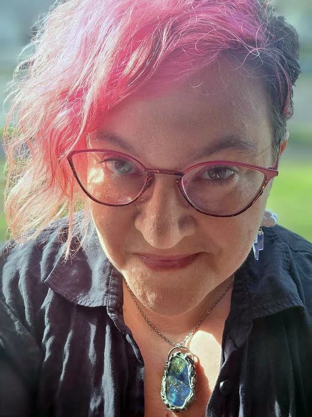 Heather FD with short pink hair, wearing large pink glasses, a black shirt, and a large gemstone necklace, smiling outdoors.