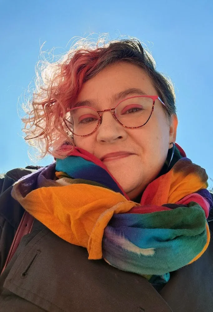Heather FD with short, curly pink hair and pink-framed glasses smiles while wearing a multicolored scarf and a dark jacket against a clear blue sky.