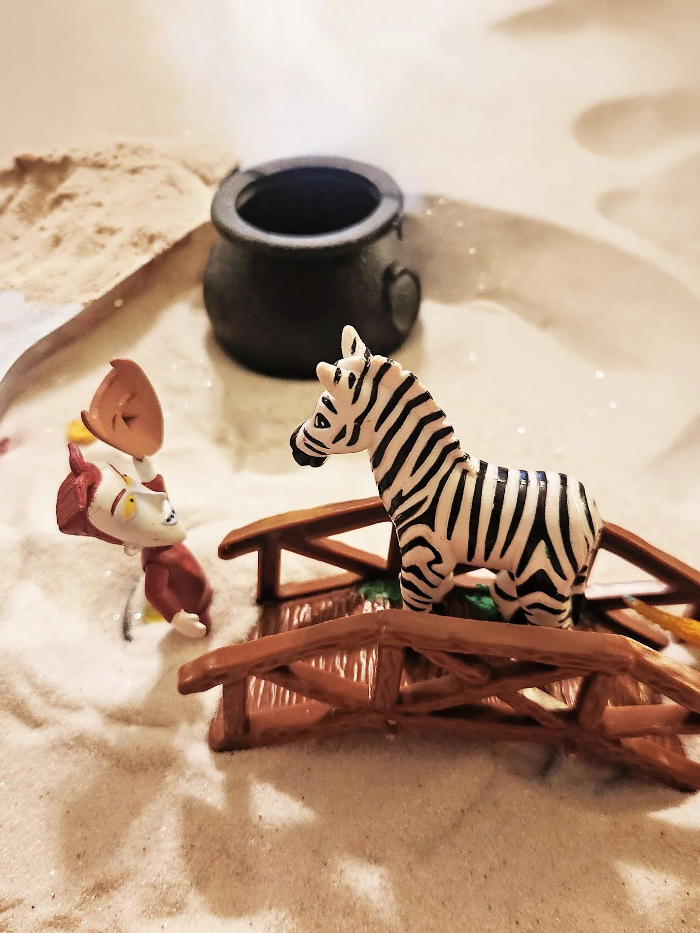 A miniature play therapy scene of a zebra figurine and a small clown figurine in a sandy environment with a small wooden fence, a black vessel, and possibly a miniature tree or structure in the background.