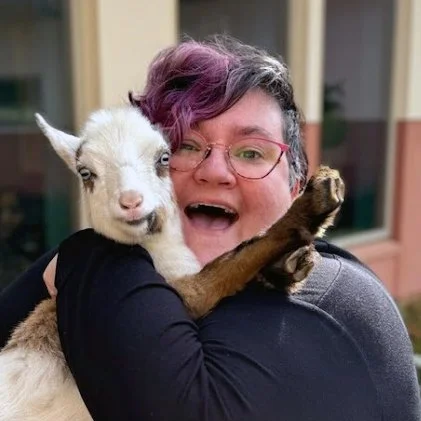 Heather FD with short purple hair, glasses, smiling, hugging a baby goat outside.