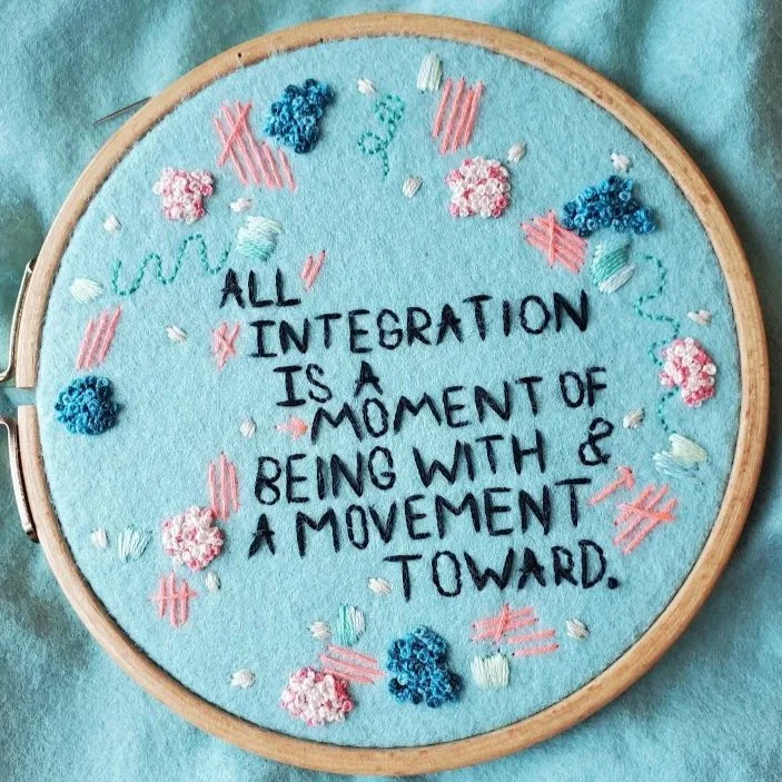 Embroidery hoop with a light blue fabric background displaying an embroidered quote. The quote reads: "All INTEGRATION is a MOMENT of BEING WITH & a MOVEMENT TOWARD."