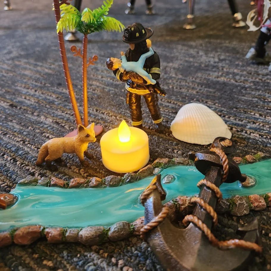Miniature play therapy scene of a firefighter figurine holding a plastic toy dog, next to a small LED candle, a lion figurine, a palm tree, a seashell, and a small river made of plastic, with some rocks and a wooden toy boat with a rope.
