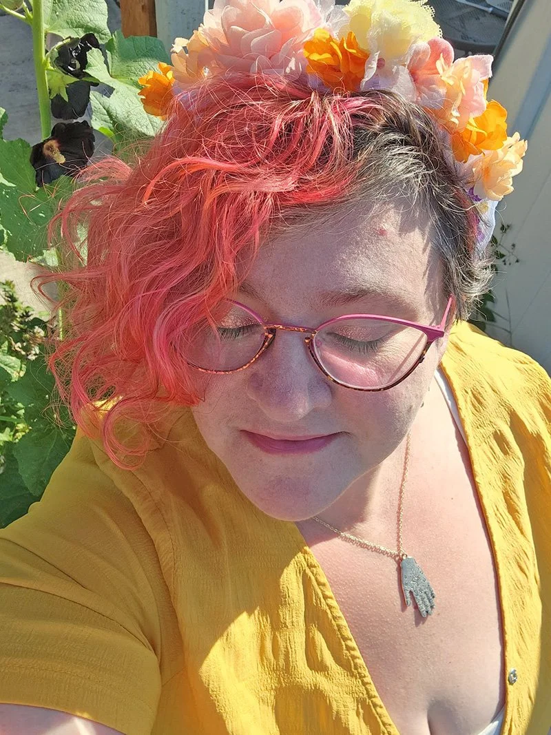 Heather DD with pink curly hair, wearing pink glasses, a floral headband with pink, yellow, and white flowers, a yellow top, and a necklace with a hand-shaped pendant. She is outdoors in the sunlight with green plants in the background.