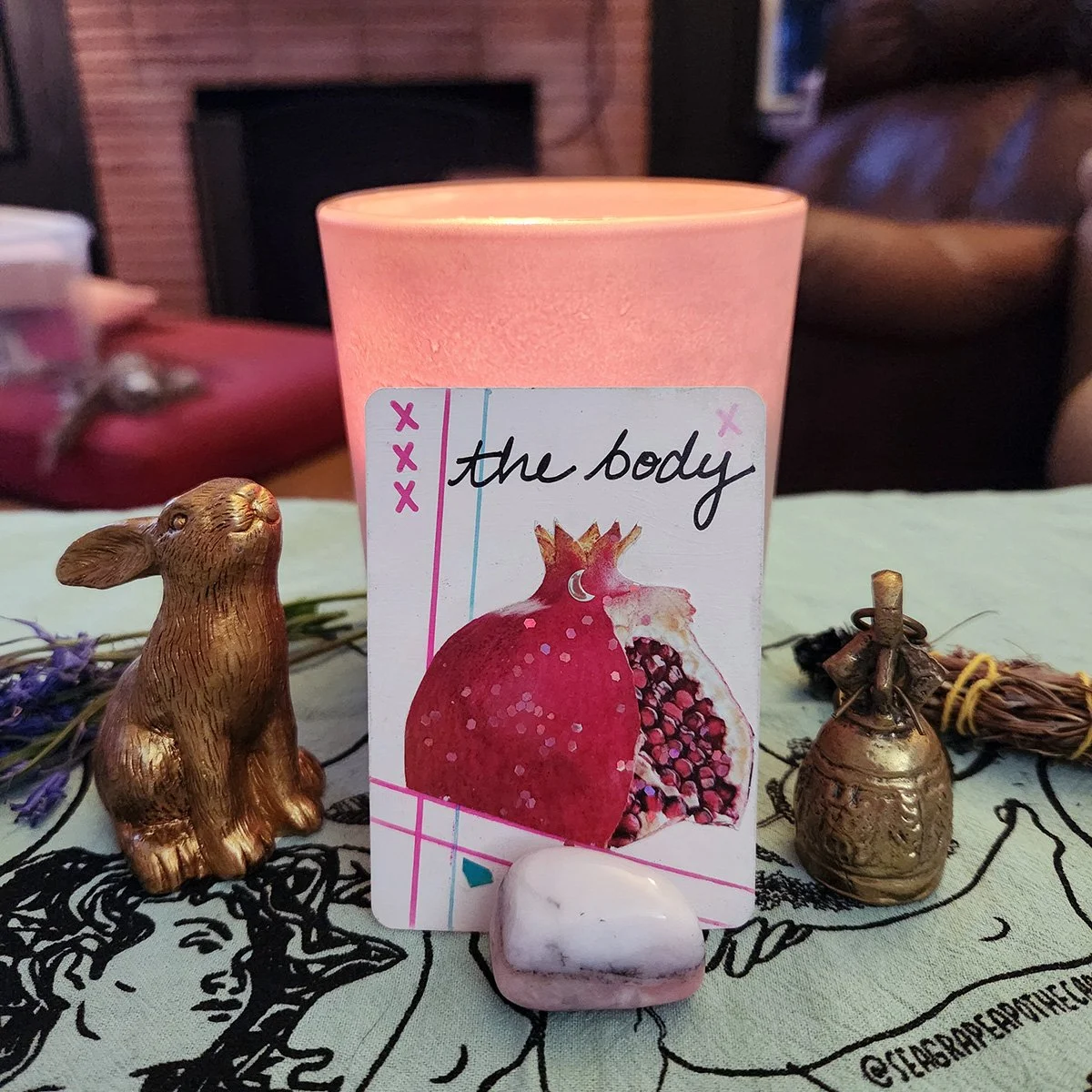 A lit pink candle, a small gold rabbit figurine, a card with an illustration of a pomegranate and the words 'the body', and a small white stone are placed on a decorated surface with floral and artistic patterns.
