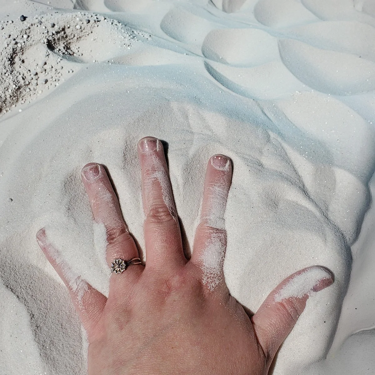 A hand with a ring on the ring finger pressing into white sand, creating a depression. The sand is sparkling and appears fine and soft.