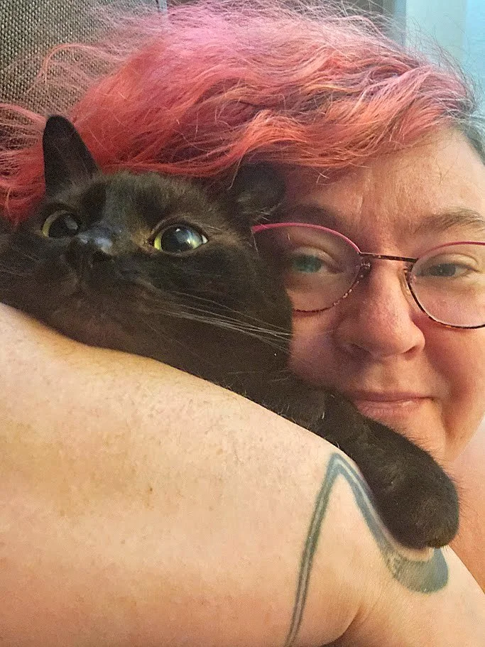 Heather FD with pink hair holding a black cat close, both looking at the camera, with the person's face partially visible and glasses, and the cat resting on their arm.