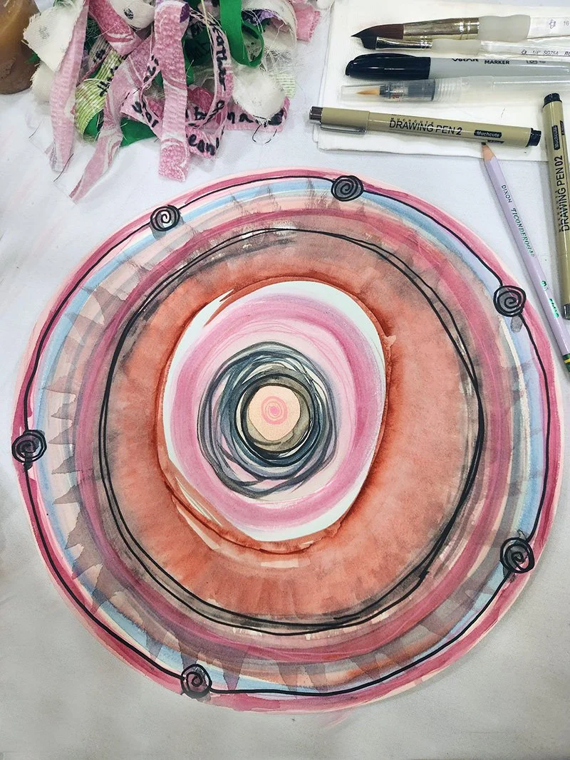 A circular watercolor painting with multiple concentric rings in shades of pink, orange, brown, and blue, bordered by black lines and spiral accents. Art supplies, including markers, pens, and fabric scraps, are visible around the painting on a white surface.