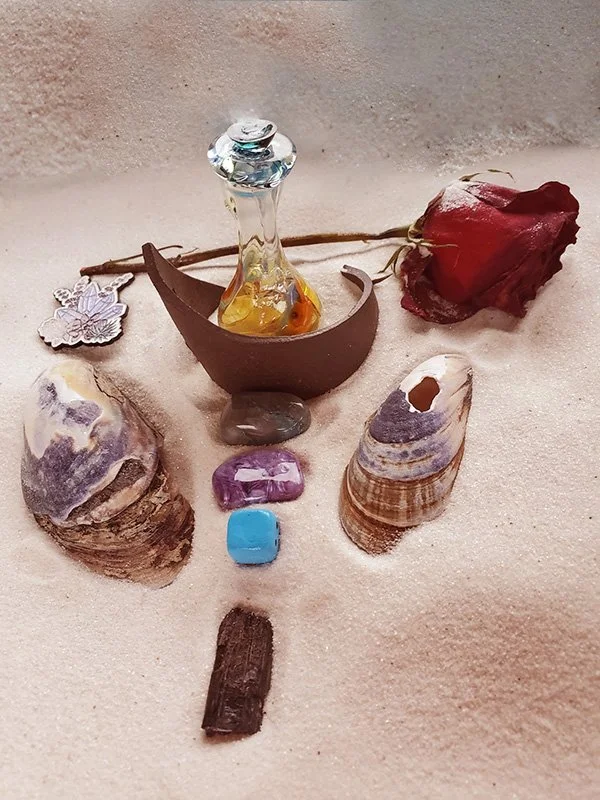 Various objects arranged on sand, including a glass bottle with liquid, shells, stones, a red dried flower, a piece of wood, a silver leaf pendant, and a small brown container.