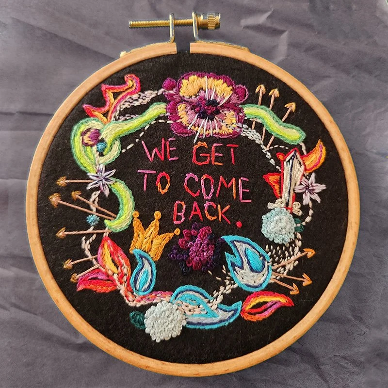 Embroidery hoop with colorful floral and leaf designs surrounding the phrase "We get to come back" stitched in pink and red on a black fabric background.