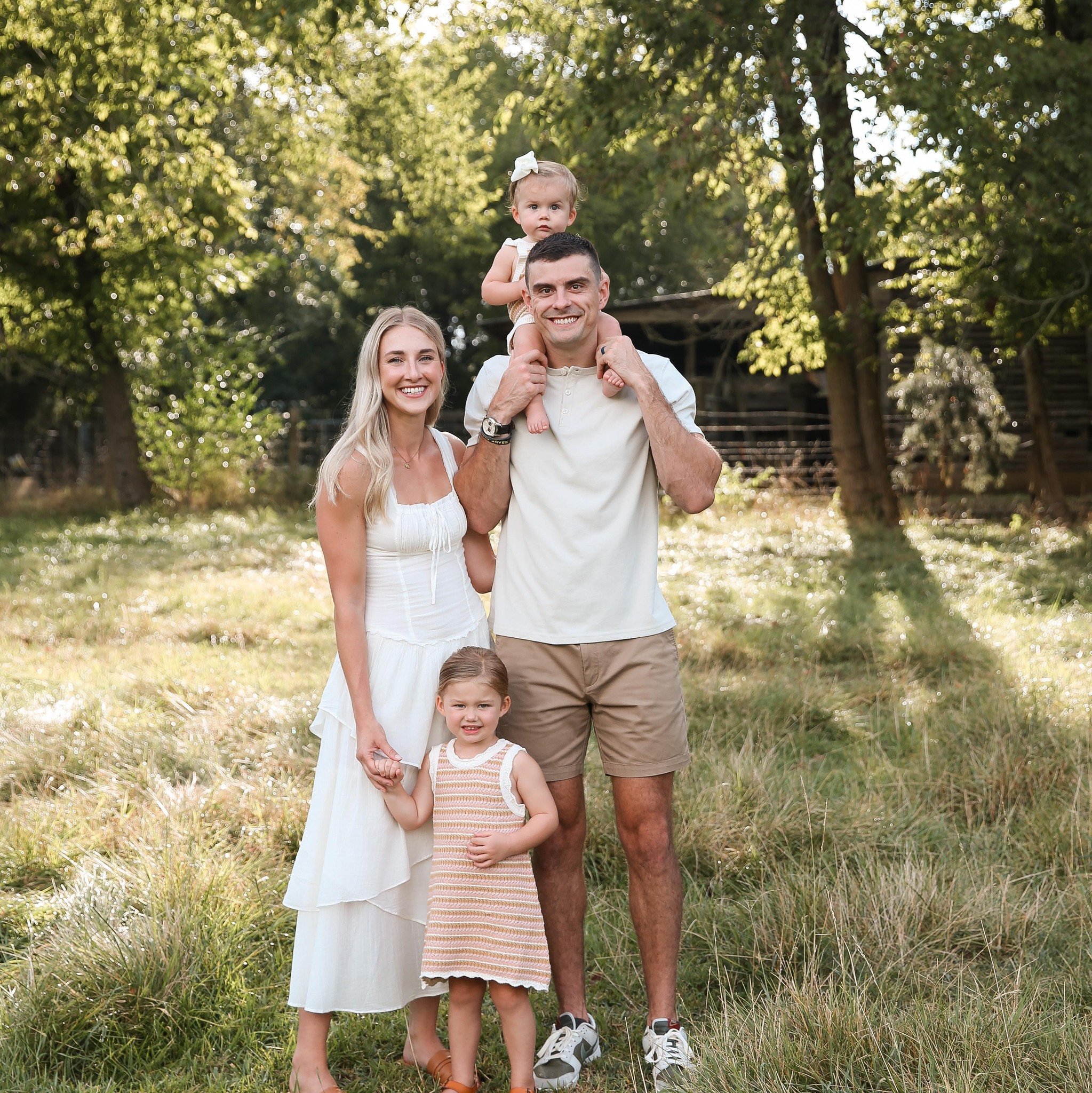 A happy family of four outdoors with trees in the background, showing a man carrying a young girl on his shoulders, a woman smiling beside him, and a young girl standing in front holding their hands.